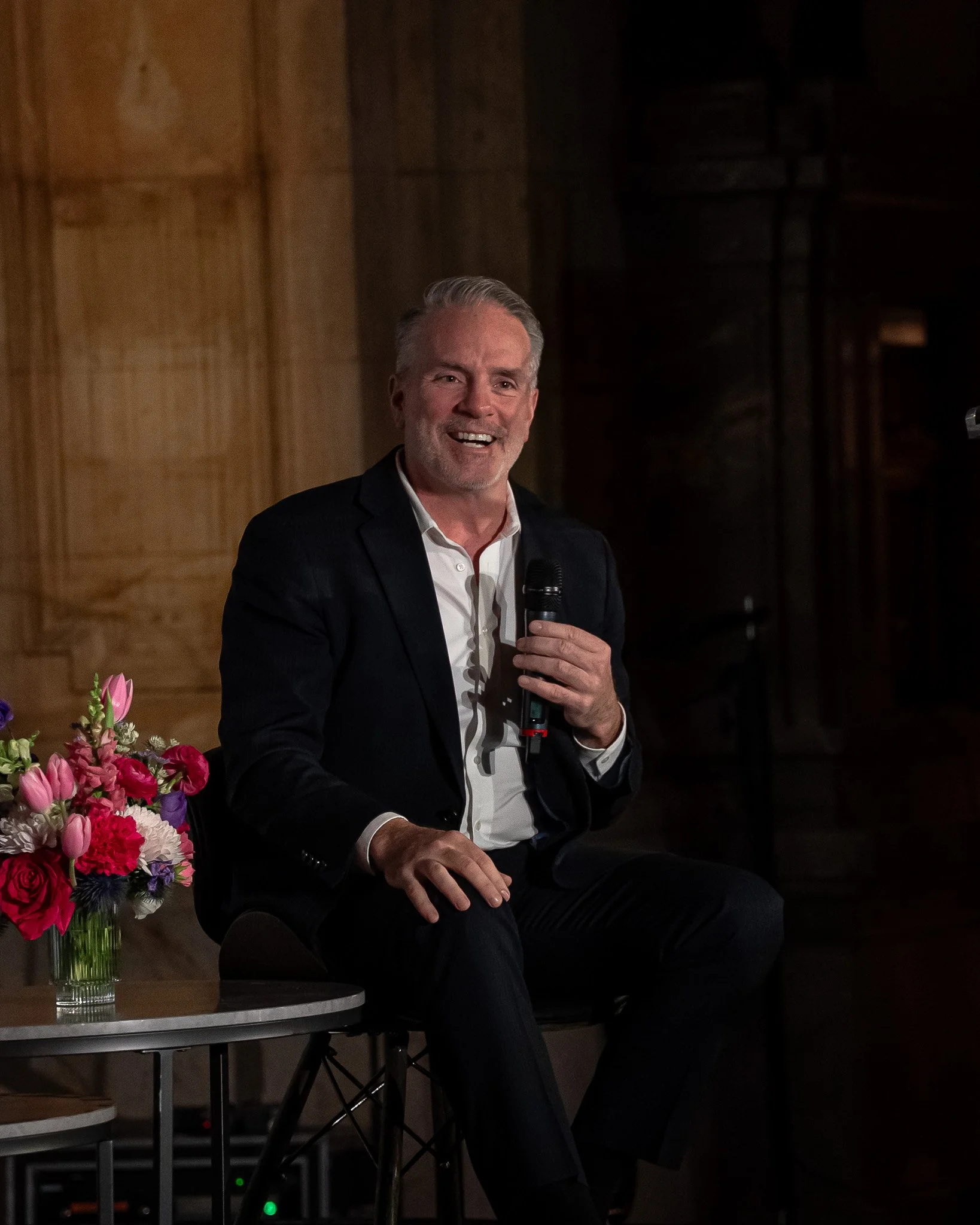 A man in a black suit and white shirt sitting on a chair holding a microphone, smiling during a speaking event, with a floral arrangement on a table beside him.