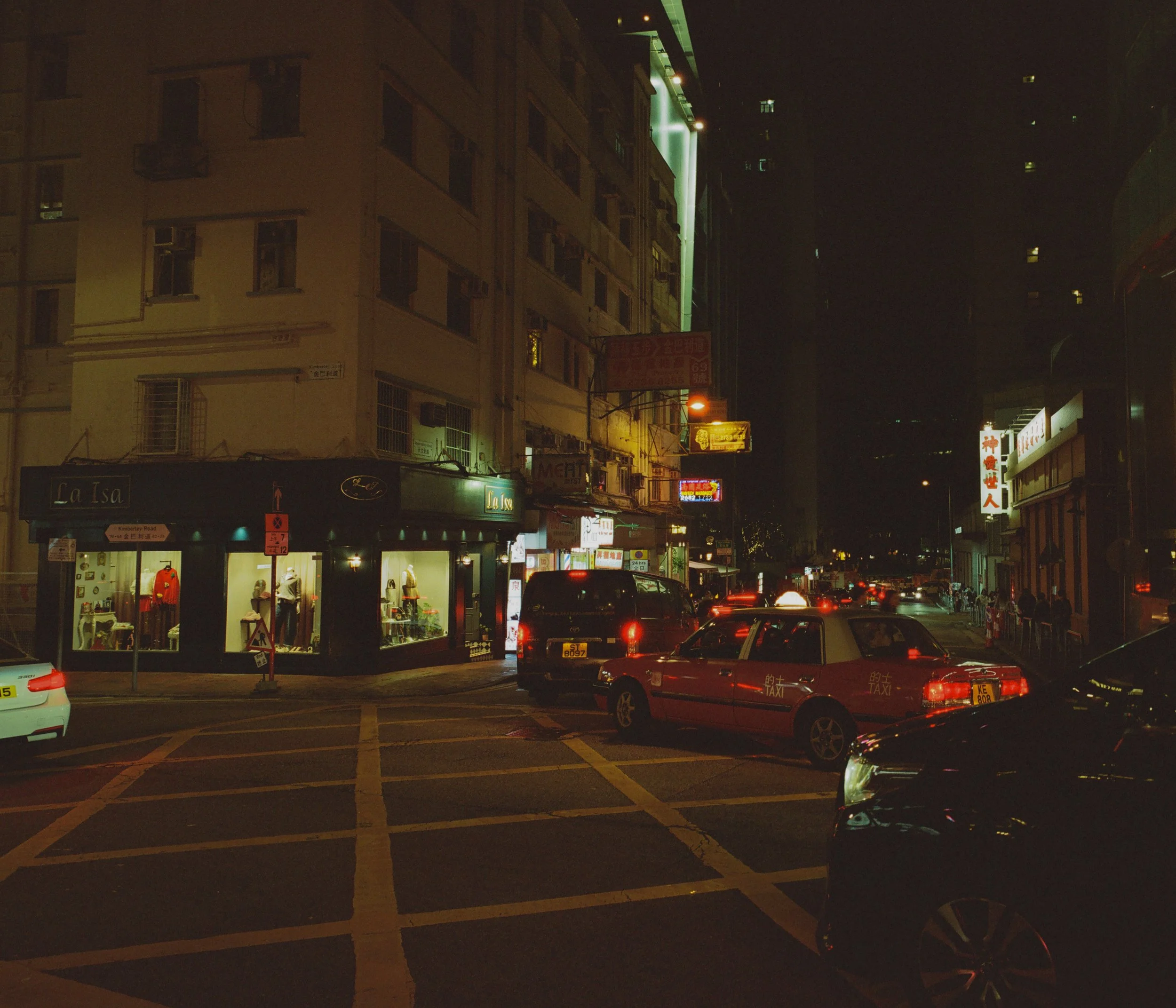 Night street scene in an urban area, illuminated by streetlights and store signs. Several cars are parked and moving along the road, with storefronts and signs visible on the sides of the street.