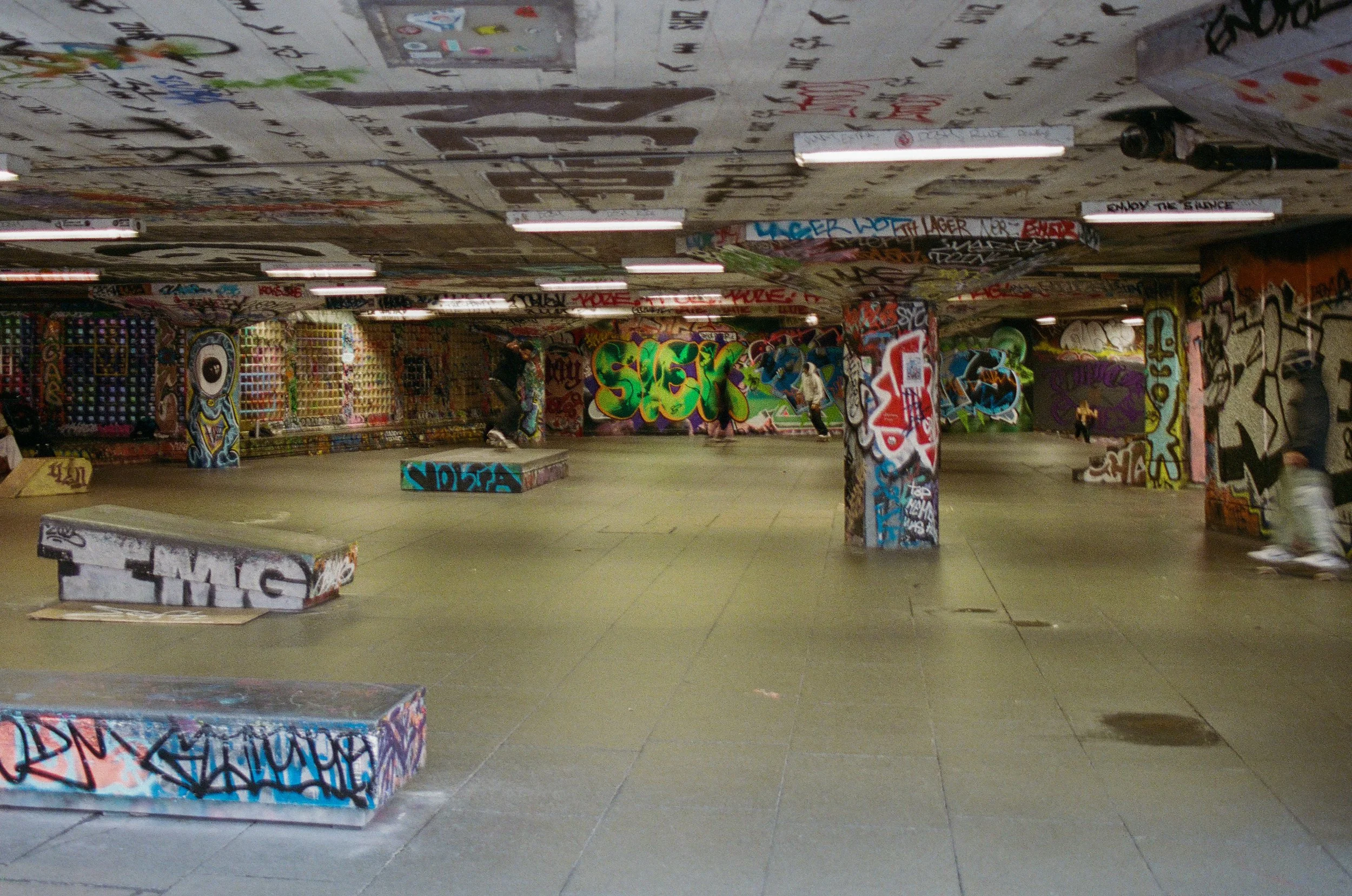 Underground skate park with graffiti-covered walls and pillars, featuring ramps and skateboards, with people skating and relaxing.
