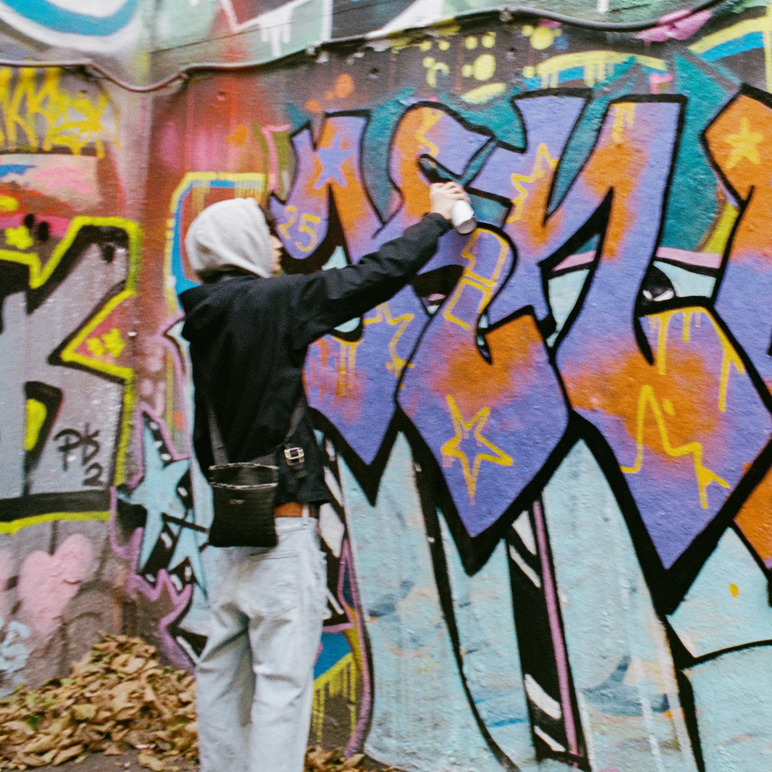 Person wearing a gray hoodie and black jacket spray painting graffiti on a colorful wall.