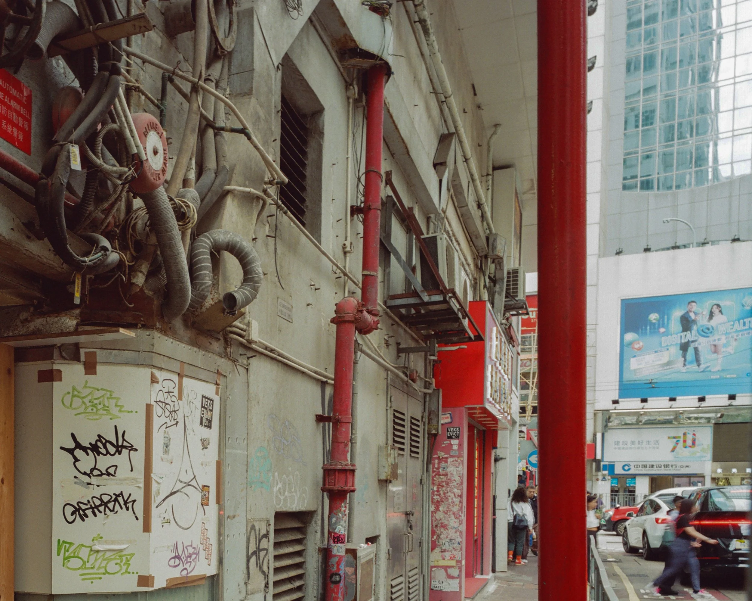 A busy urban street scene with pedestrians walking past buildings and parked cars. The buildings have various commercial signs, billboards, and advertisements. Bright red structural poles and a mix of modern and older architecture are visible.