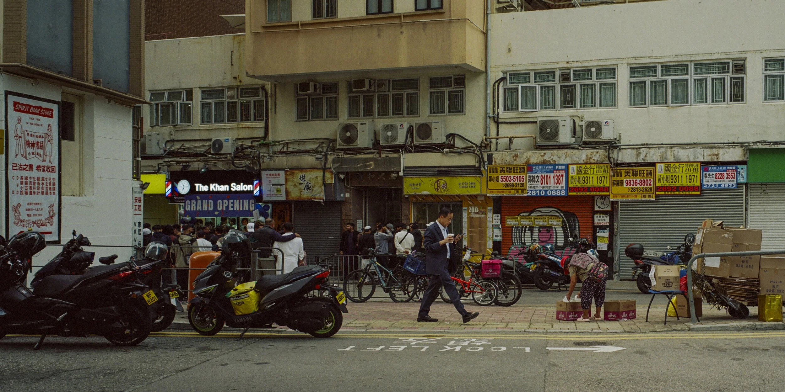 A busy street scene in an Asian city with people, motorcycles, bicycles, and storefronts, including a barber shop announcing a grand opening.