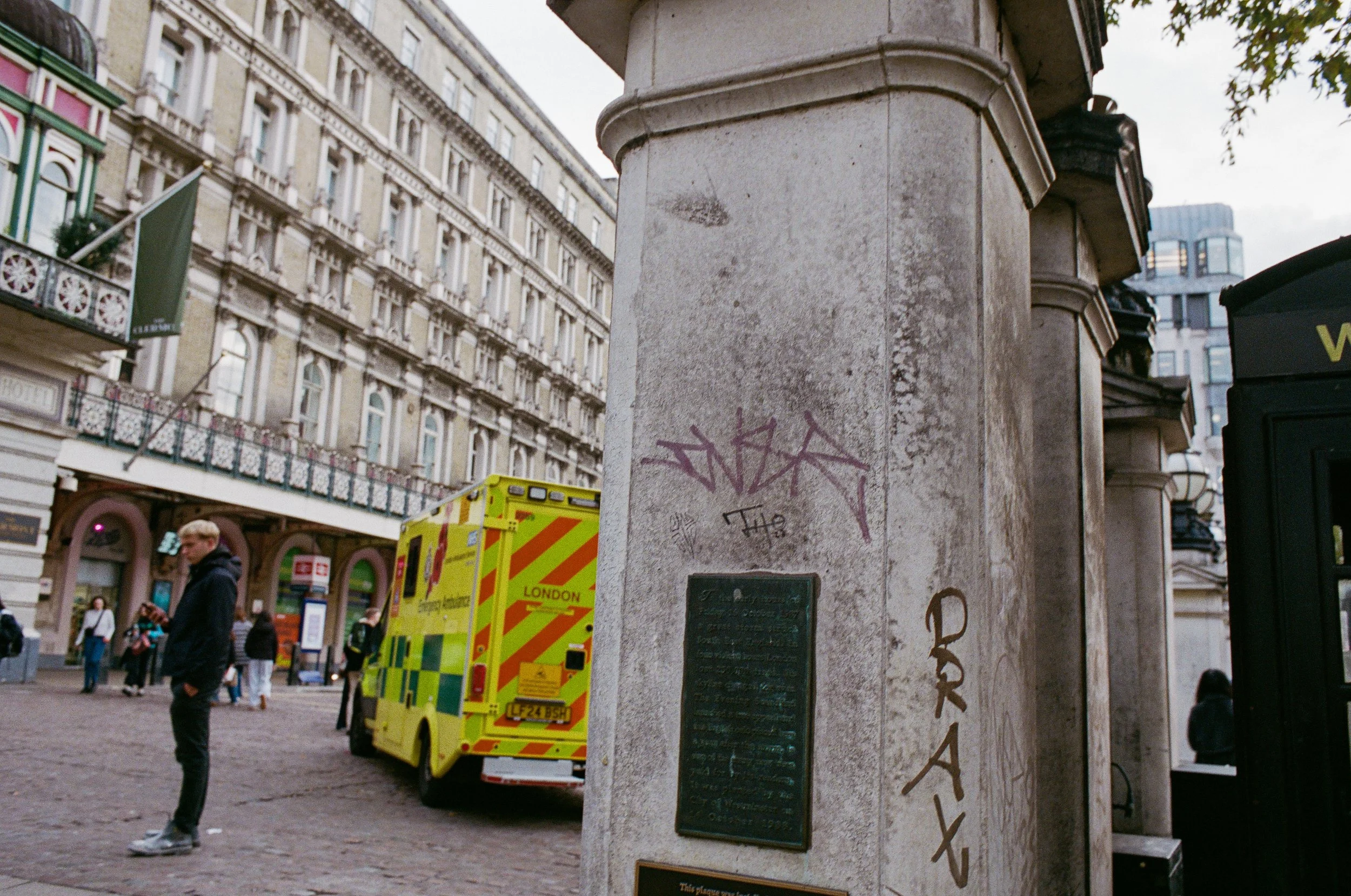 A city street scene featuring a man in a black hoodie standing on cobblestone pavement, with a yellow ambulance parked nearby. In the foreground, a stone monument with graffiti and plaques is visible. The background shows historic multi-story buildin