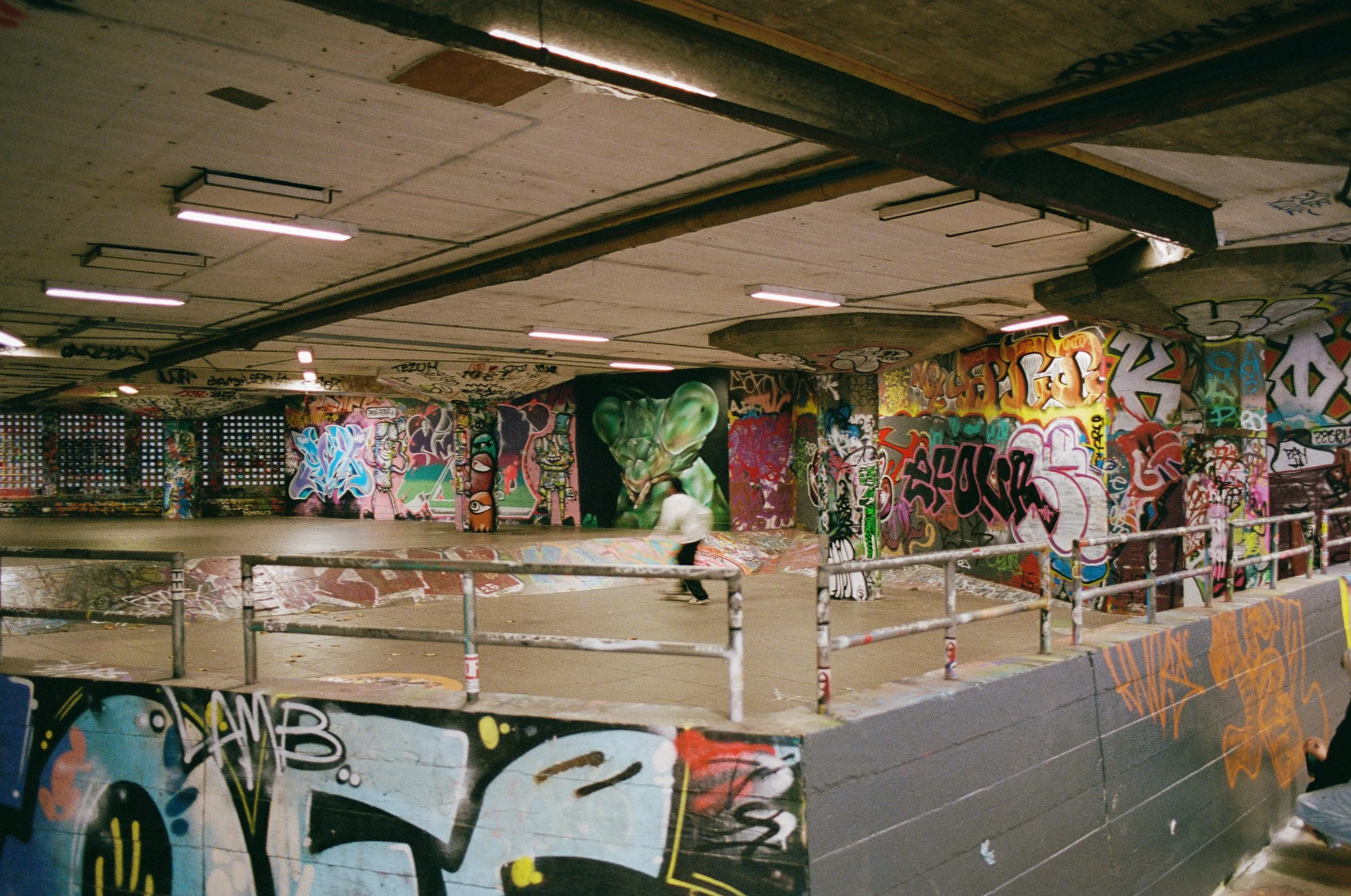 An underground skate park with graffiti-covered walls, a skateboarder riding on a ramp, and a person sitting on the ledge in the foreground.