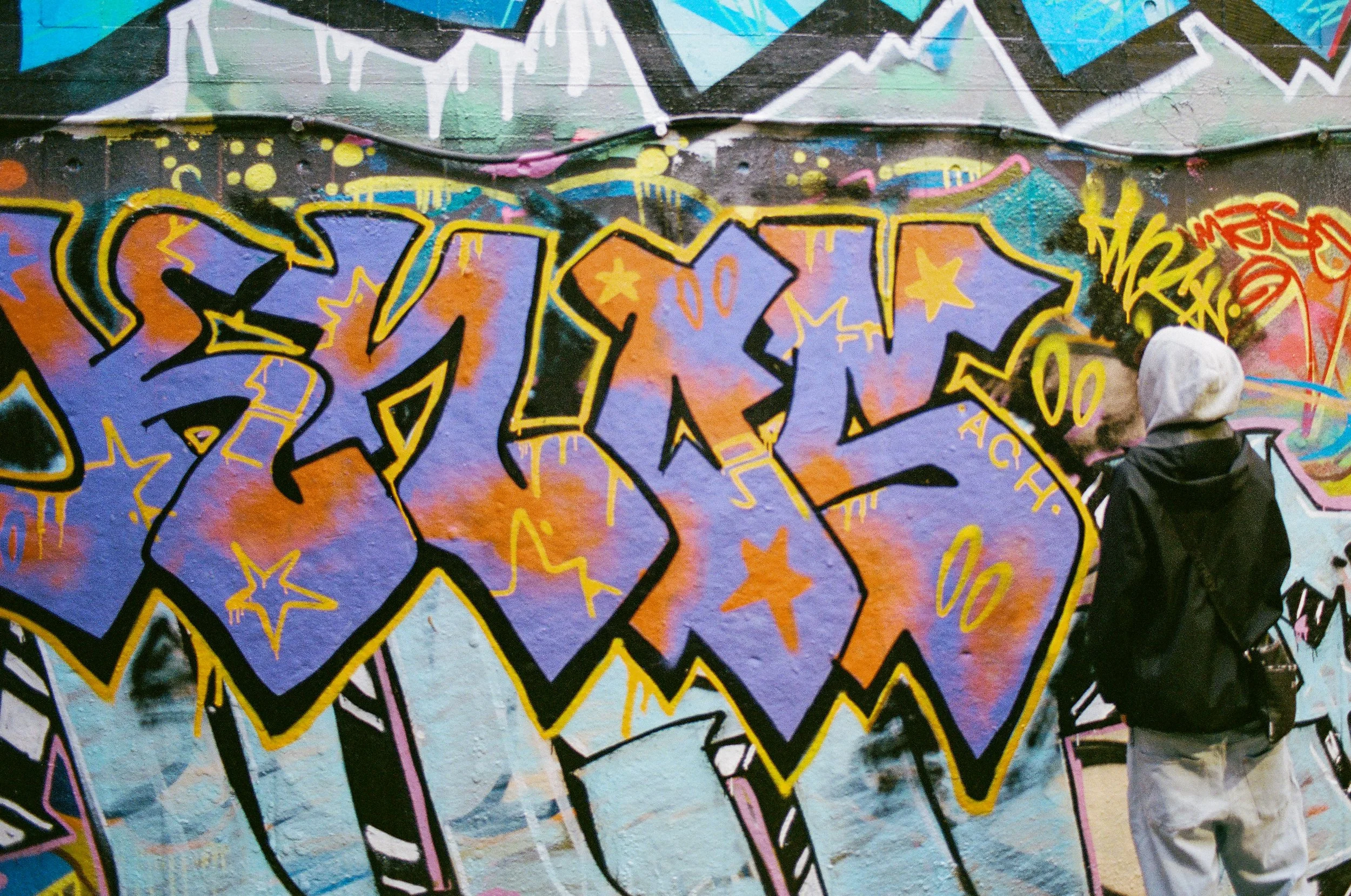 A person with a hoodie and black jacket standing in front of a colorful graffiti wall with large purple and orange bubble letters and yellow accents, stars, and abstract shapes.