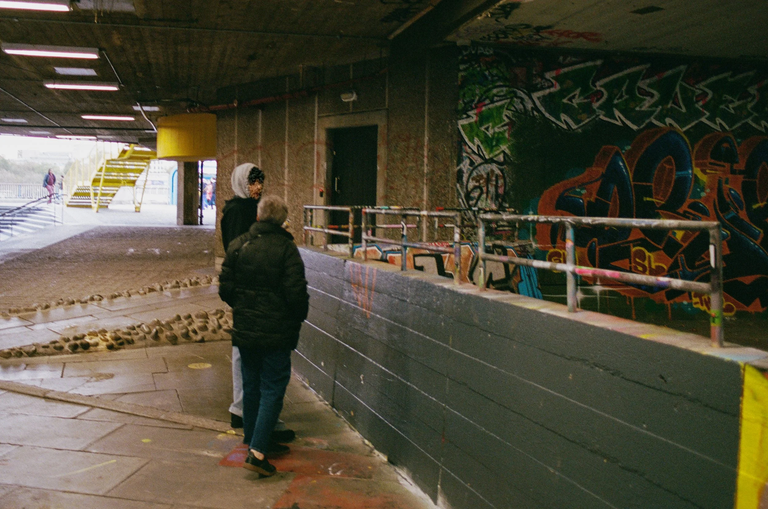 Two people stand on a sidewalk under an overpass with graffiti on the wall behind them, having a conversation. In the background, there is a yellow staircase leading to an outdoor area with people walking in the distance.