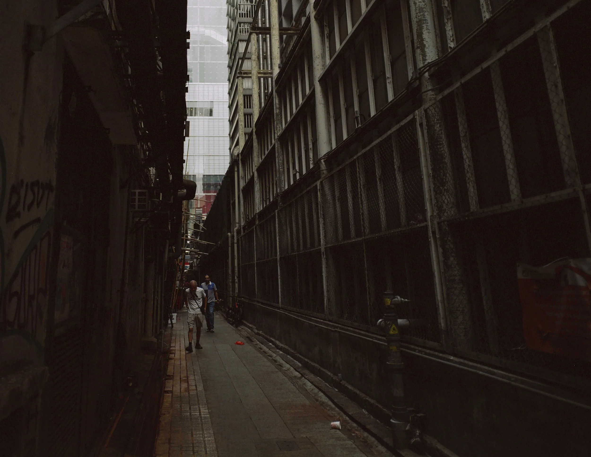 A narrow alleyway between tall buildings with scaffolding and construction fencing on the right and graffiti on the left. Three people are walking in the distance under overcast sky.