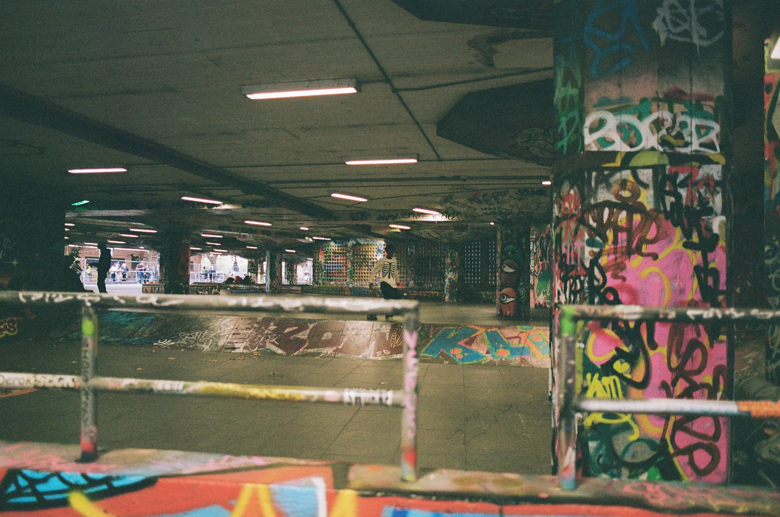 Interior view of an urban skate park with graffiti-covered walls and columns, with some skateboarders in the background.