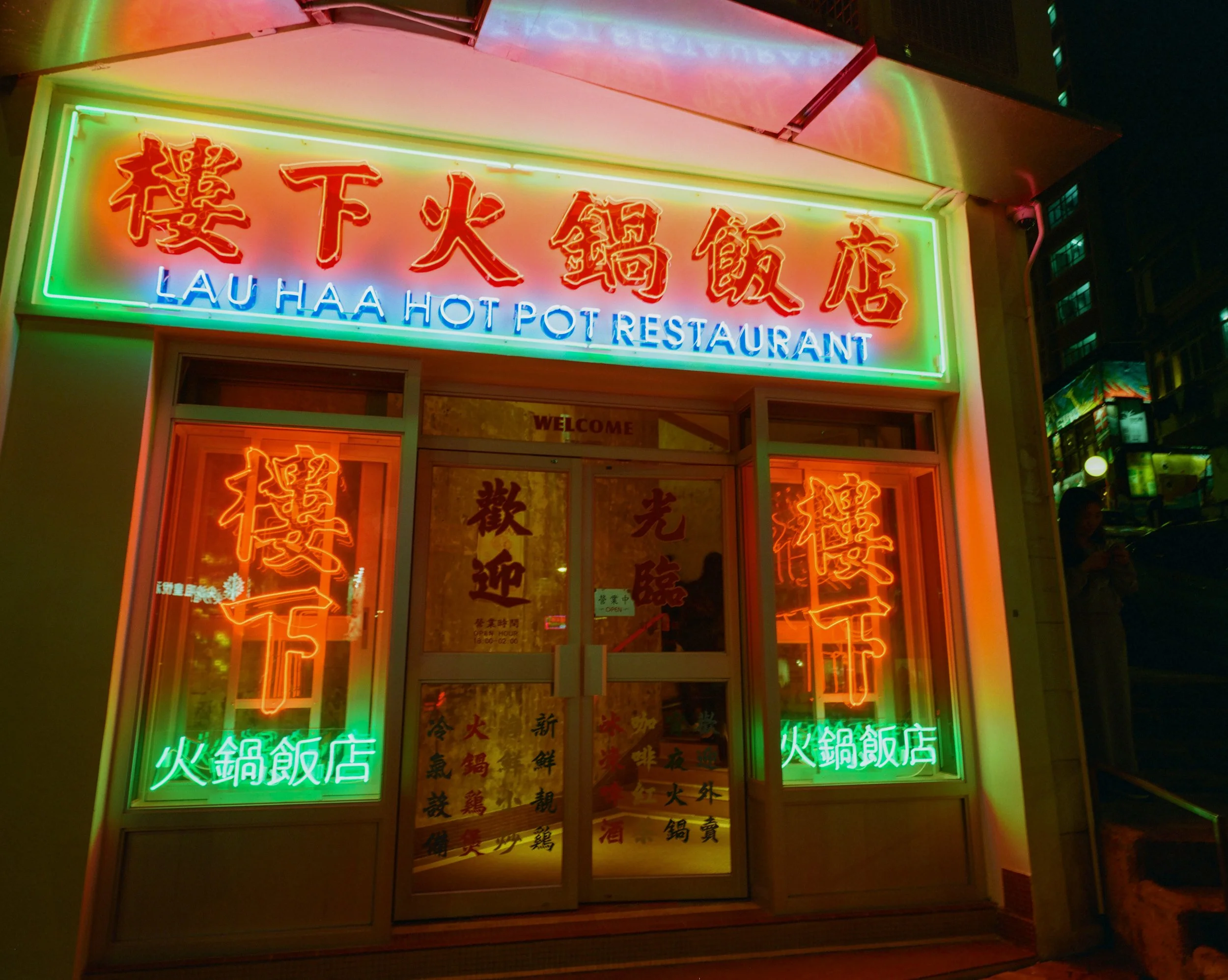 Neon-lit signs on the storefront of Lau Haa Hot Pot Restaurant, with Chinese characters and the restaurant's name in English, illuminated with red, green, and blue lights at night.