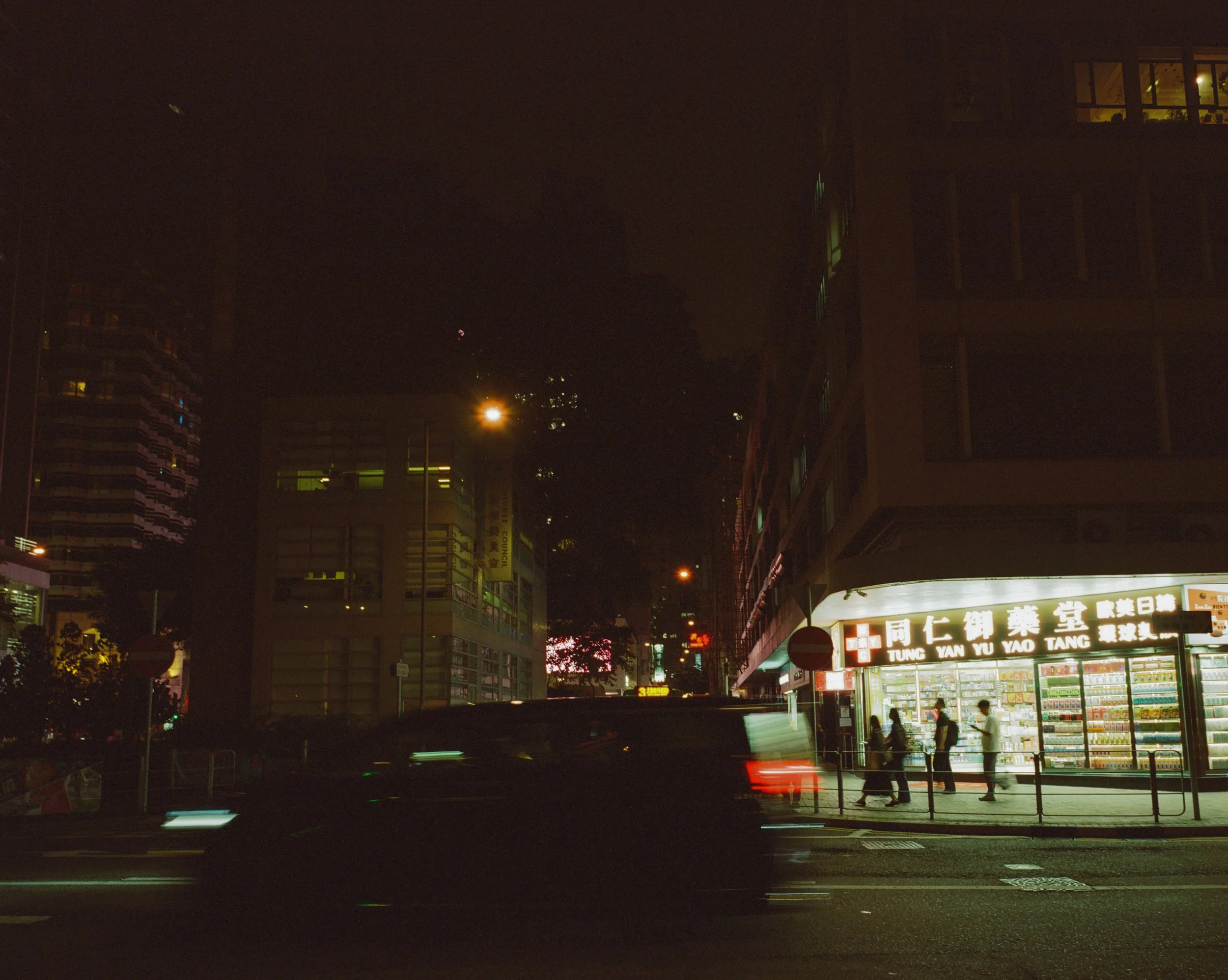 Nighttime city street scene with illuminated storefront, pedestrians, and blurred vehicle passing by.