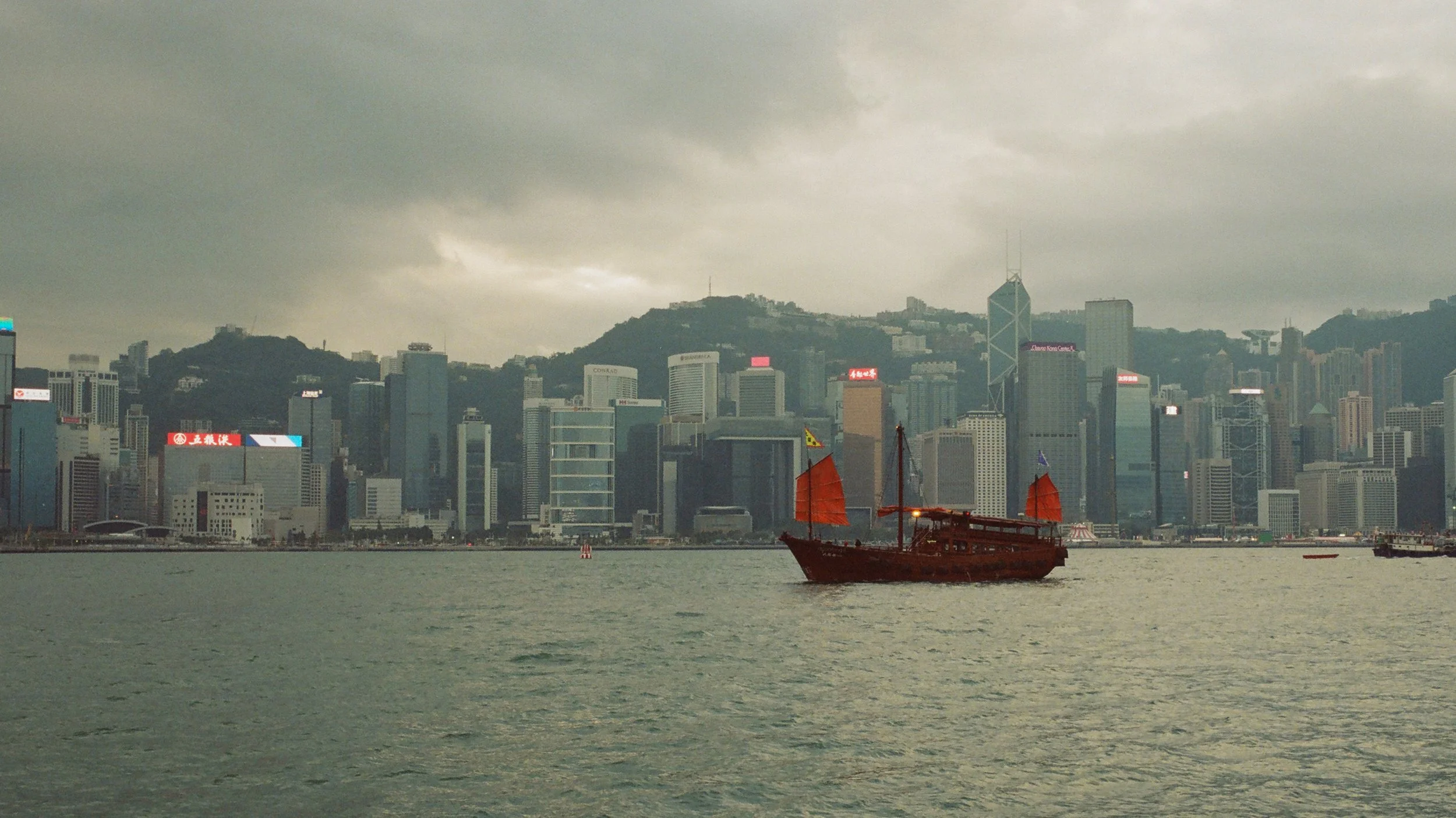 A wooden boat with red sails on a body of water with the city skyline of Hong Kong in the background, under a cloudy sky.