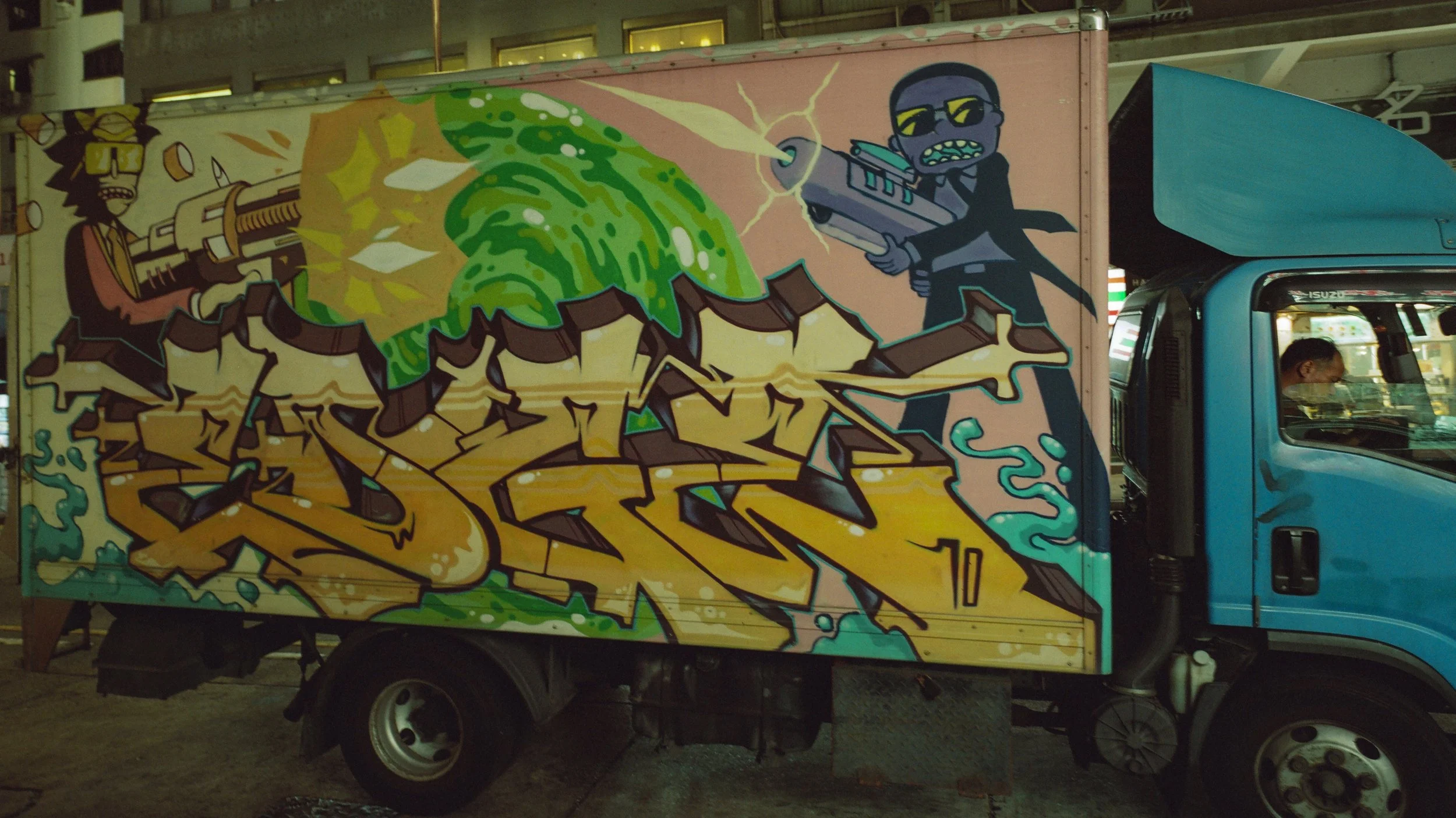 Colorful graffiti mural on the side of a blue truck, featuring cartoon characters with sunglasses and bullets, graffiti lettering in yellow and green, and a city street background.