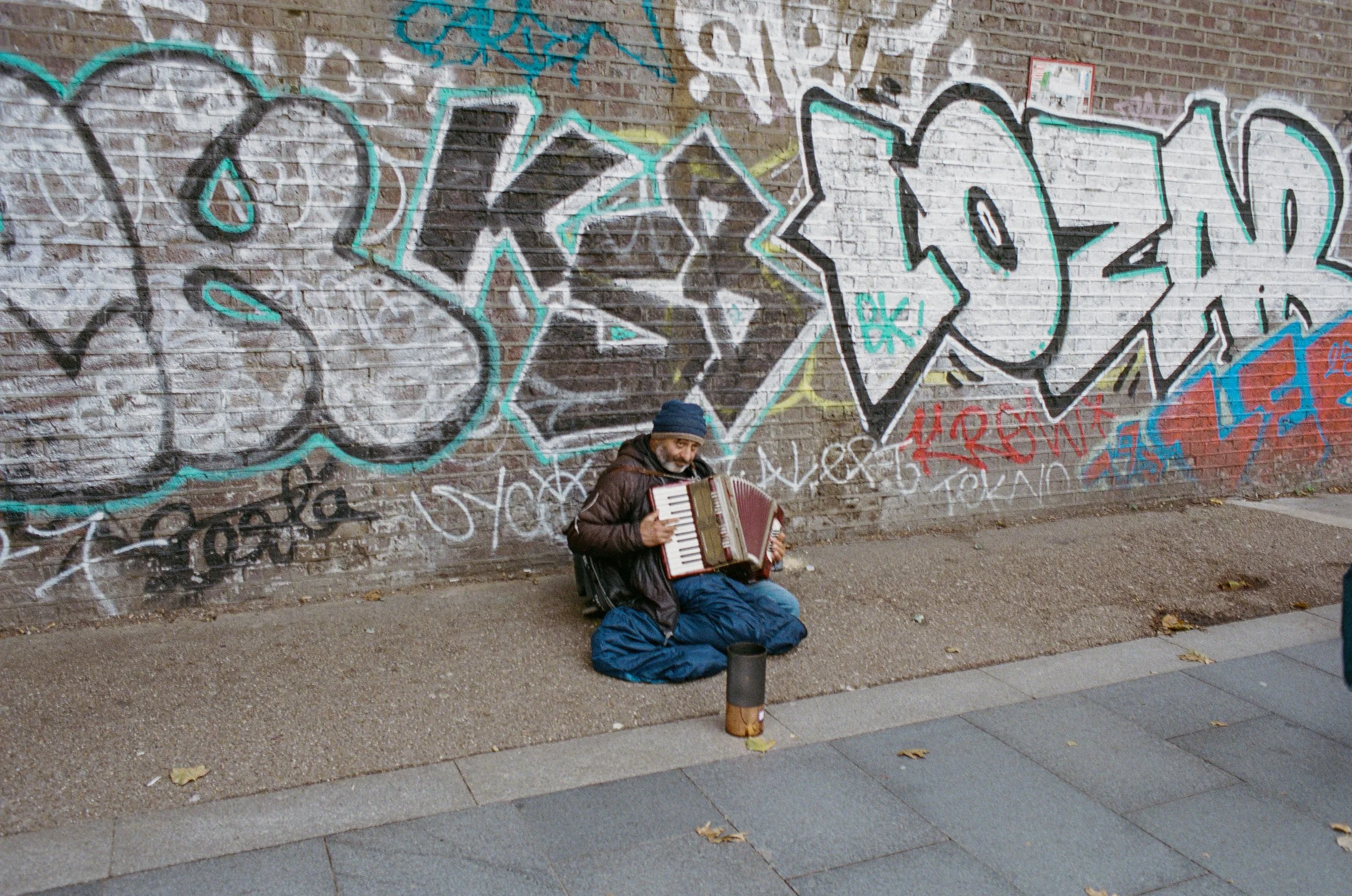 A man sitting on the sidewalk against a graffiti-covered brick wall, playing an accordion.