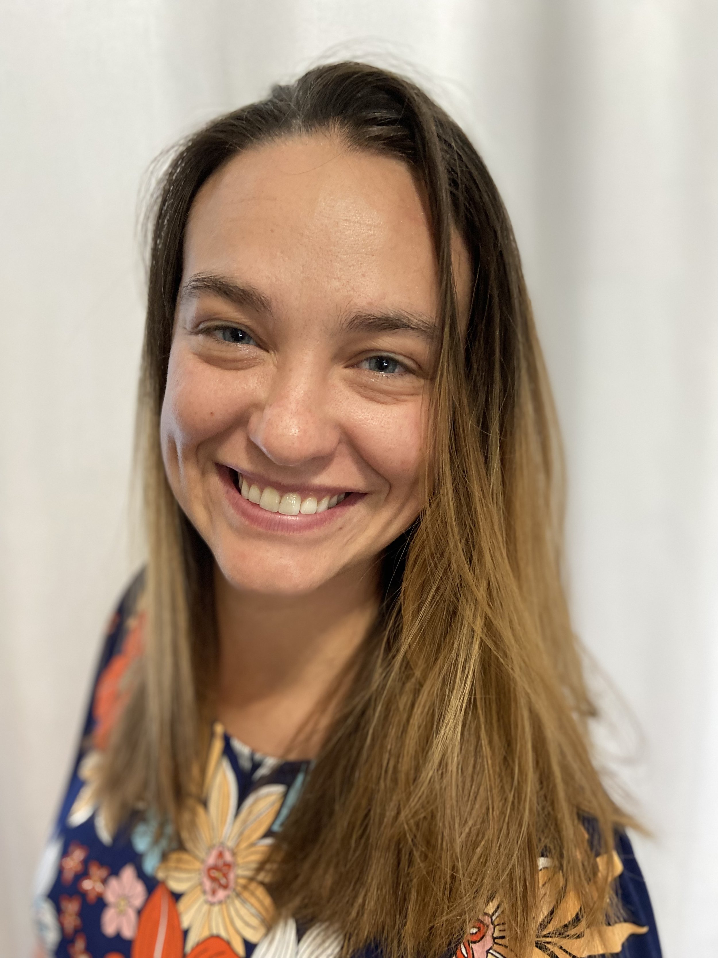 Close-up of a smiling young woman with long brown hair and blue eyes, wearing a colorful floral top, standing against a plain white background.