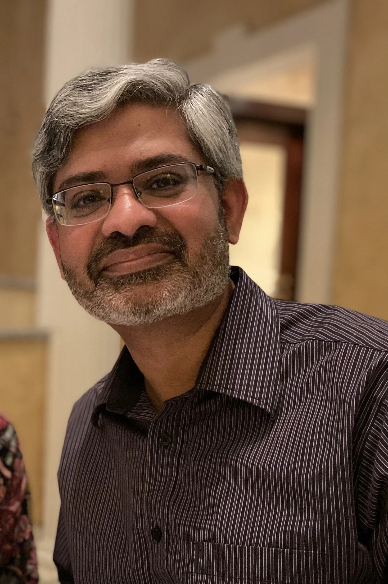 A middle-aged man with gray hair and beard, wearing glasses and a dark striped shirt, smiling at the camera indoors.