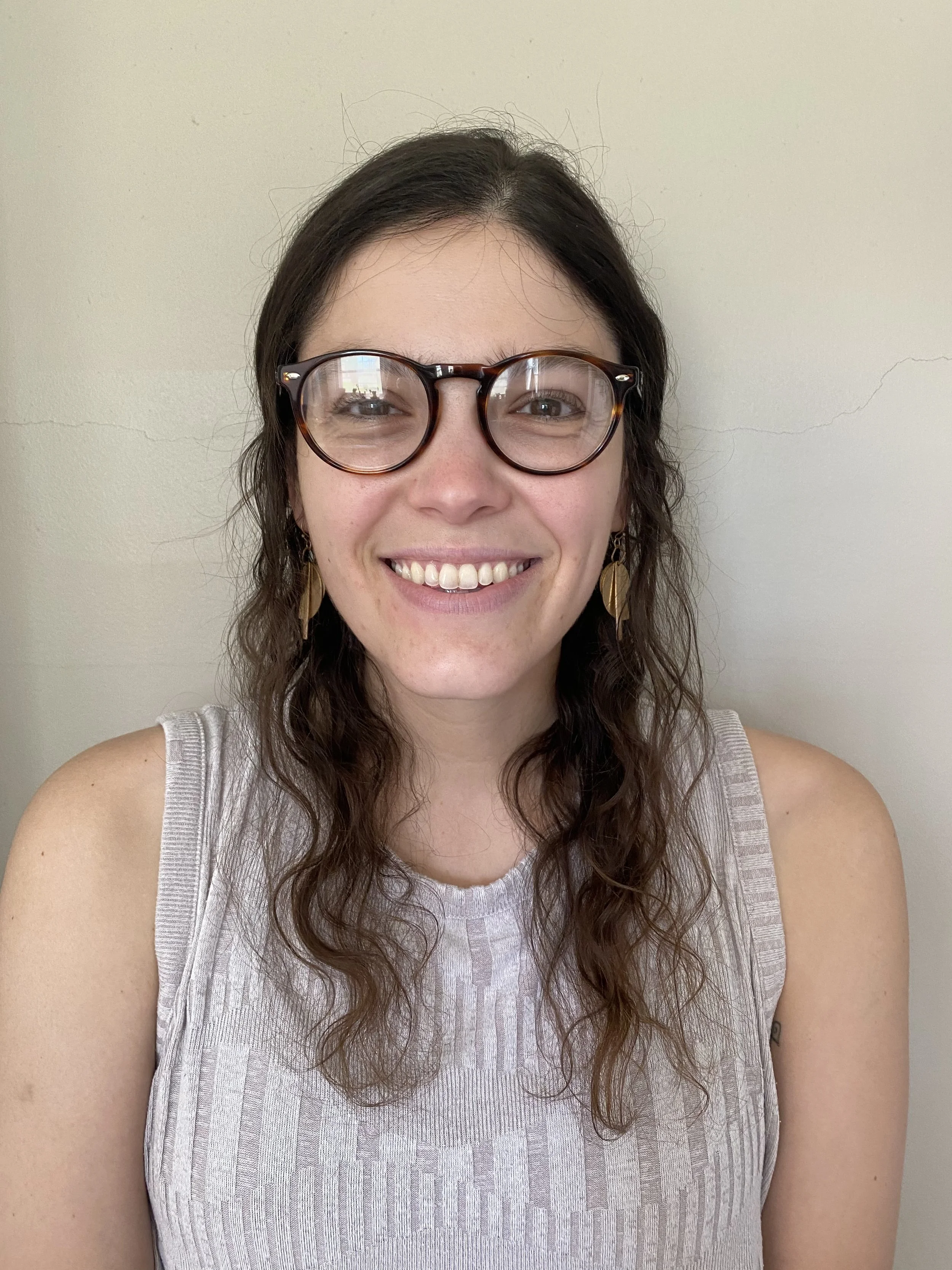 A woman with wavy brown hair and glasses smiling at the camera against a plain wall.