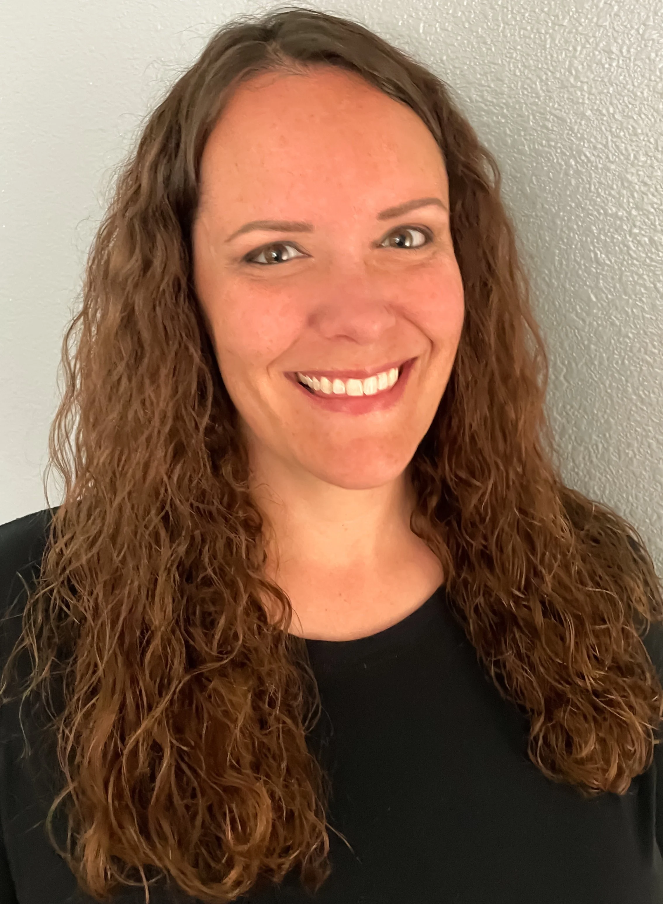 A woman with long, curly red hair smiling at the camera, wearing a black top, standing against a plain, light-colored wall.