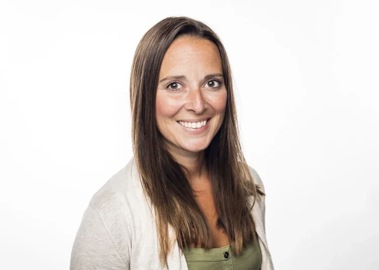 A woman with long brown hair smiling, wearing a light-colored cardigan and a green top, standing against a plain white background.