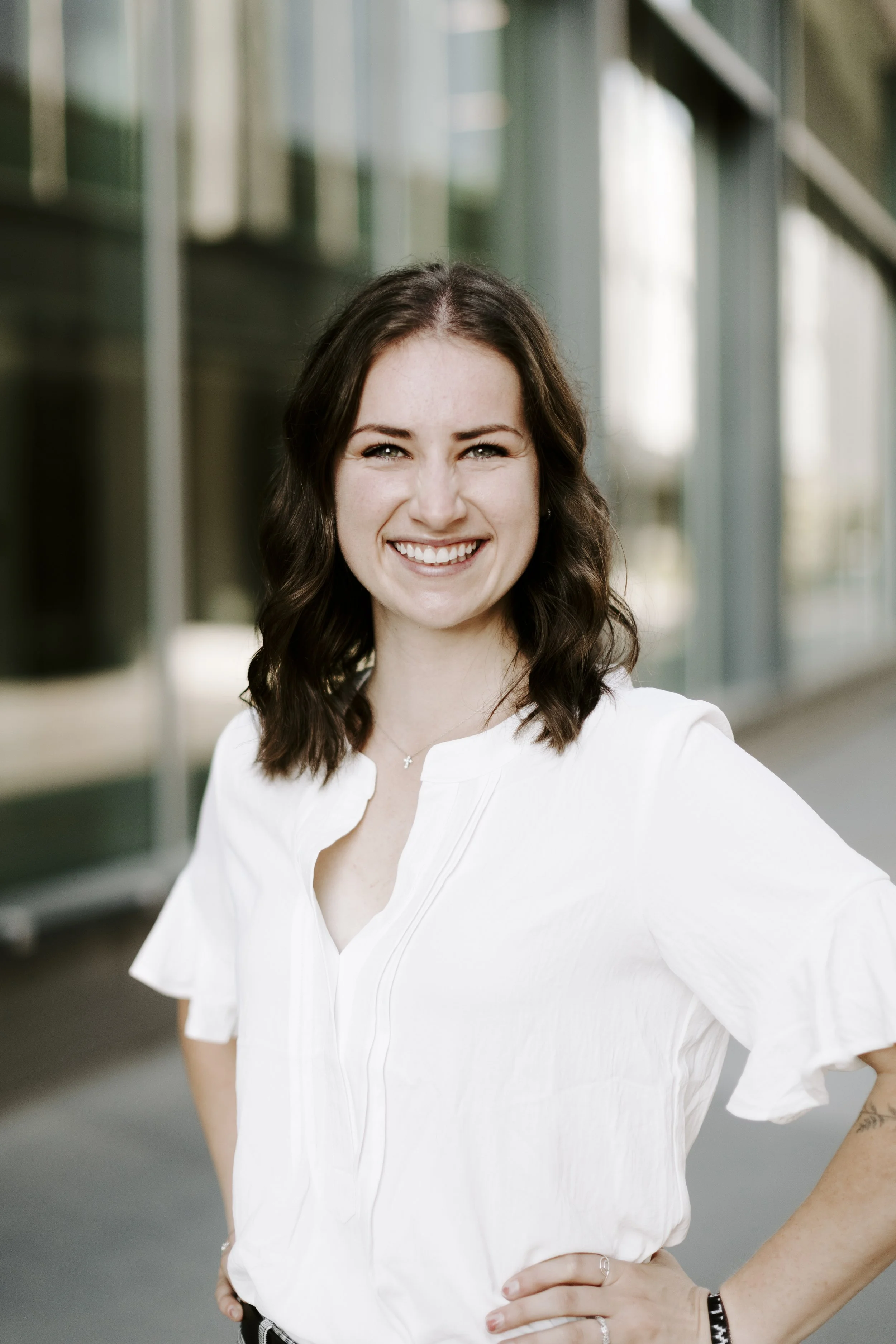 A young woman with shoulder-length dark wavy hair, smiling confidently, wearing a white blouse, standing outside with large glass windows in the background.
