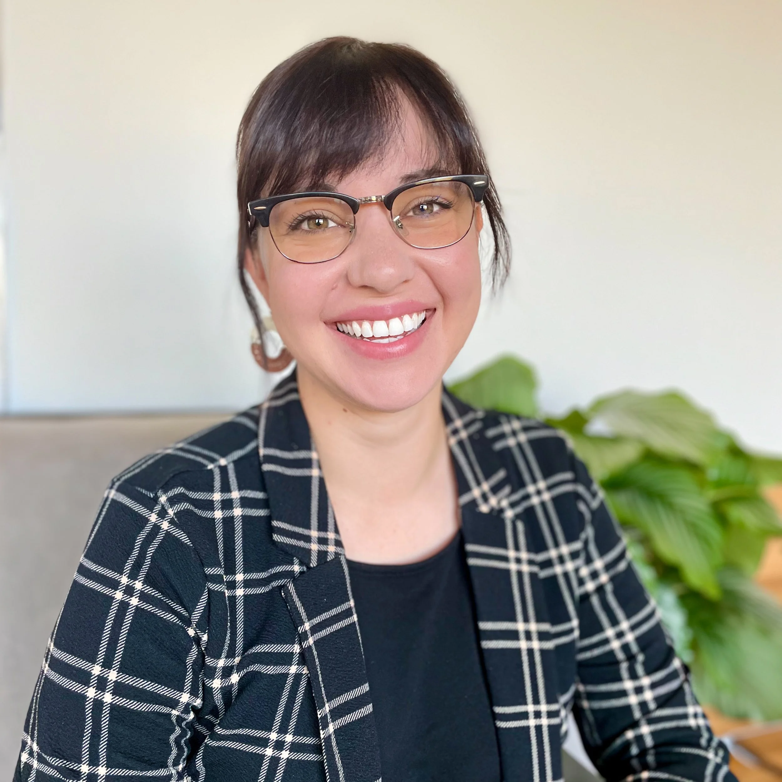 A woman with short dark hair, glasses, and a big smile, wearing a plaid blazer, sitting at a desk with a green plant in the background.