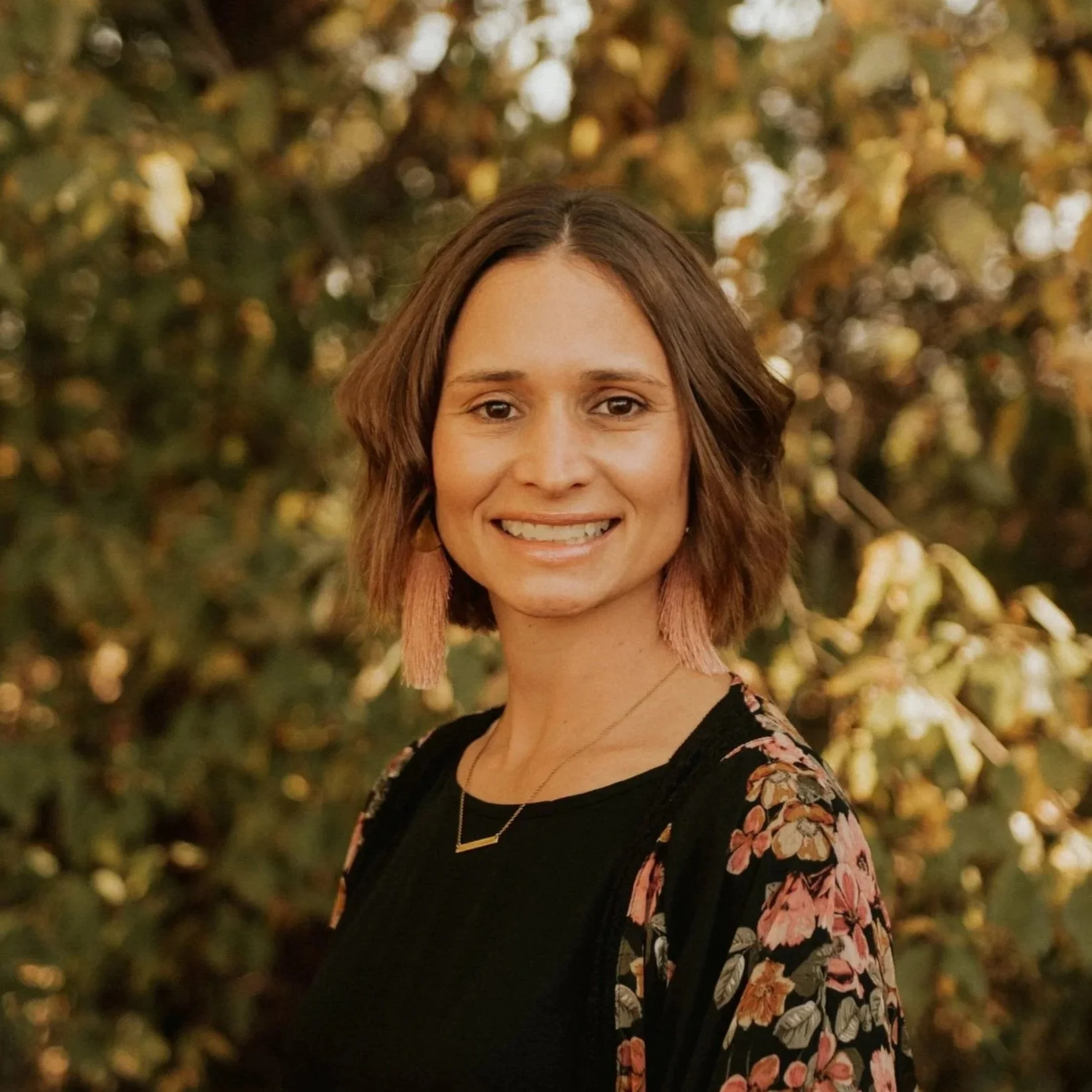 A woman smiling outdoors with a background of autumn-colored leaves, wearing a black top with floral patterns and pink tassel earrings.