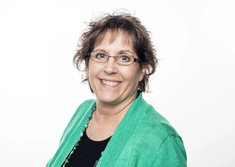A woman with short, curly brown hair, glasses, and a big smile, wearing a black top and a turquoise cardigan.