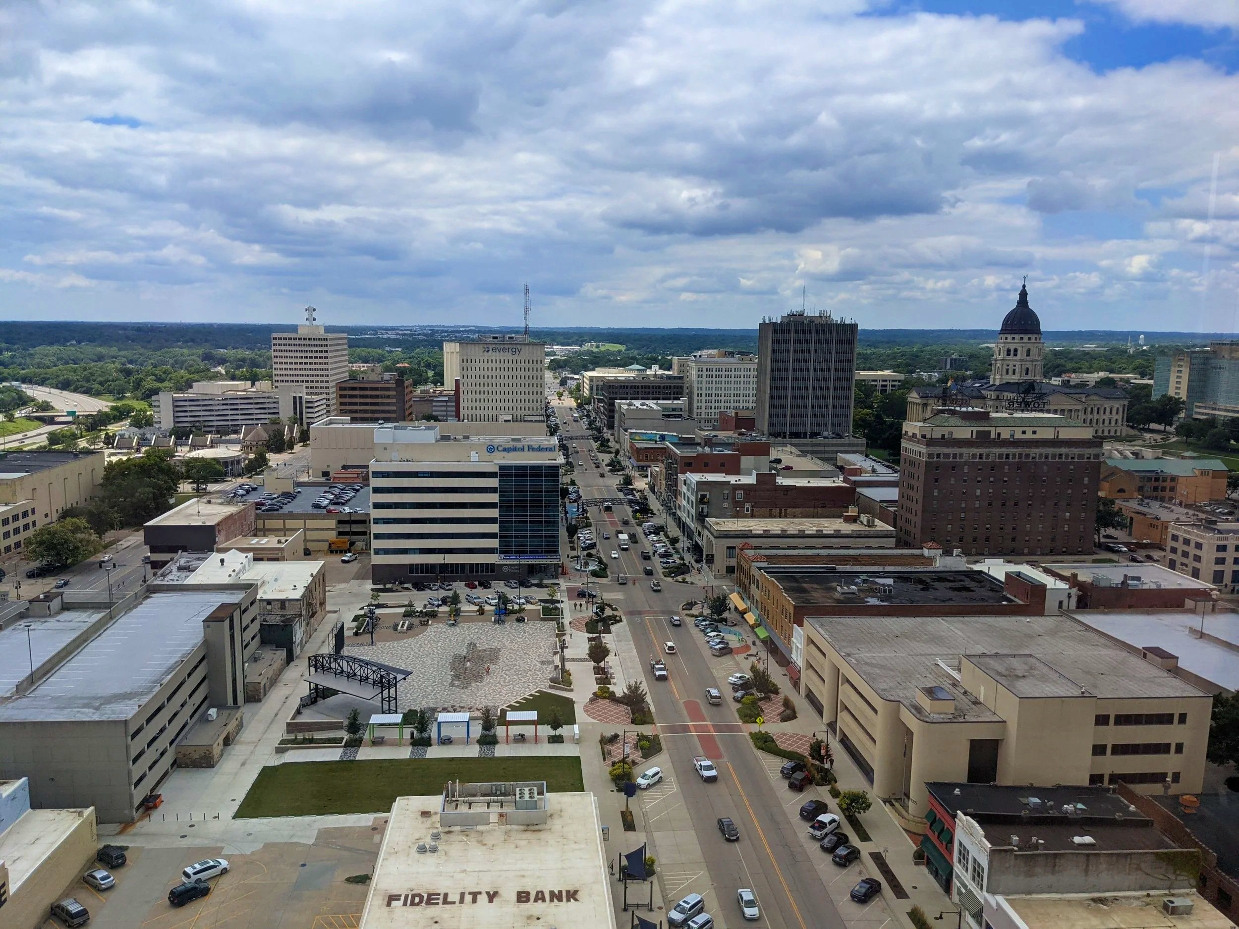 A wide aerial view of a cityscape featuring tall buildings, streets with cars, and a partly cloudy sky.