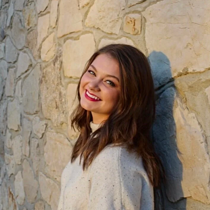 A smiling woman with long brown hair and red lipstick leaning against a stone wall outdoors during daytime.