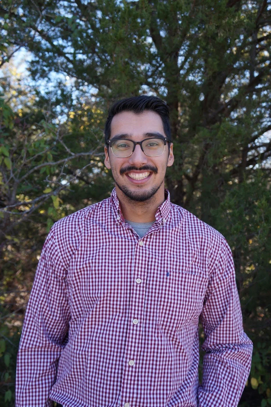 A young man with glasses, a mustache, and a goatee, smiling, wearing a red and white checkered shirt, standing outdoors in front of trees.