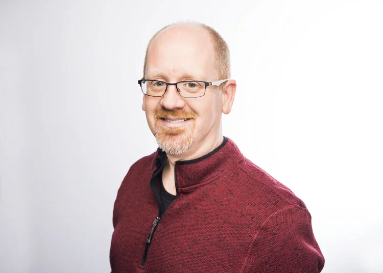 Headshot of a smiling man with glasses, short reddish hair, and a beard, wearing a maroon sweater over a black shirt against a plain white background.