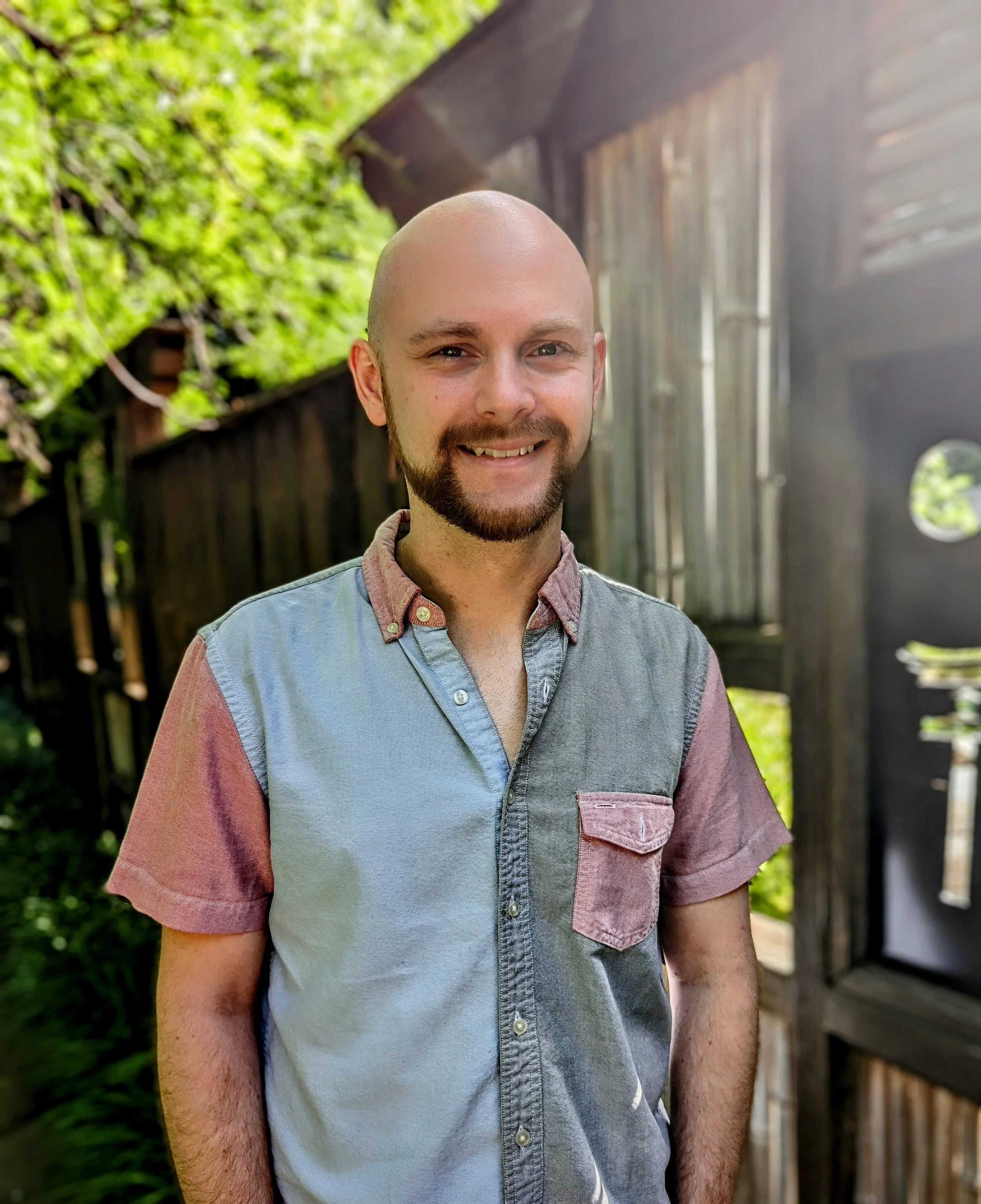 A smiling man with a beard and shaved head standing outdoors in front of a wooden building with greenery overhead.