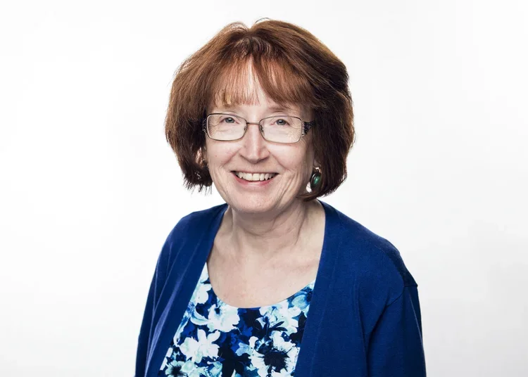 A smiling older woman with short brown hair, glasses, wearing a blue floral blouse and a dark blue cardigan against a white background.
