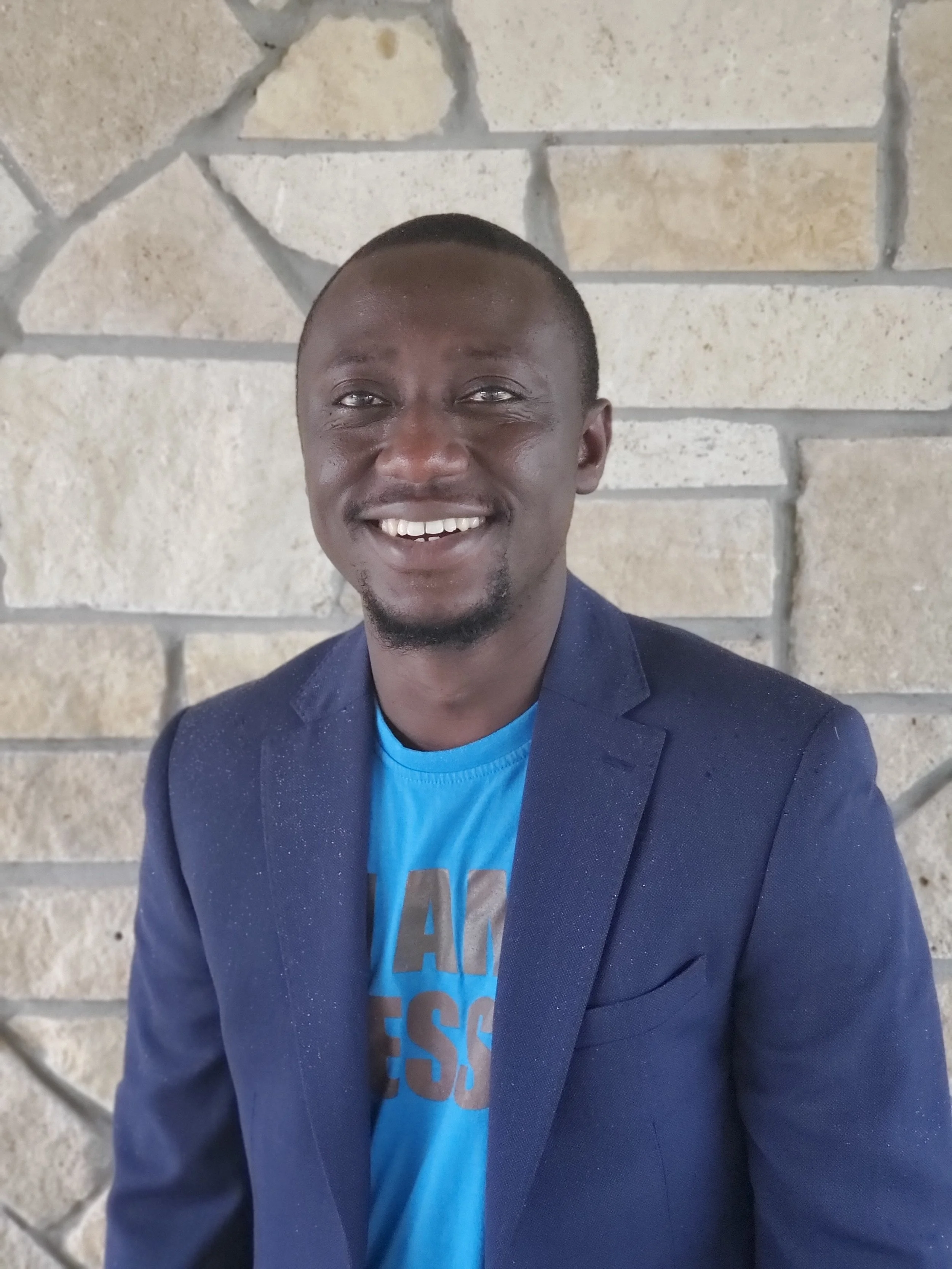 A smiling man with short hair and a beard, wearing a blue blazer over a blue t-shirt, standing in front of a stone wall.