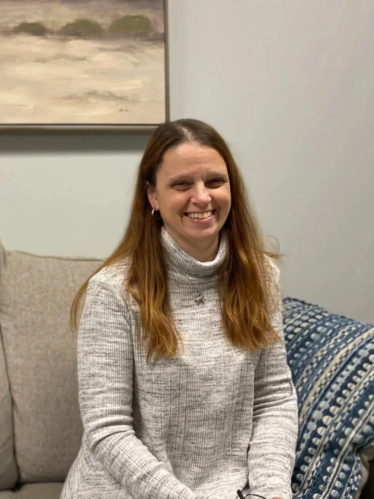 A woman with long red hair smiling, sitting on a beige couch, wearing a light gray turtleneck sweater in a living room.