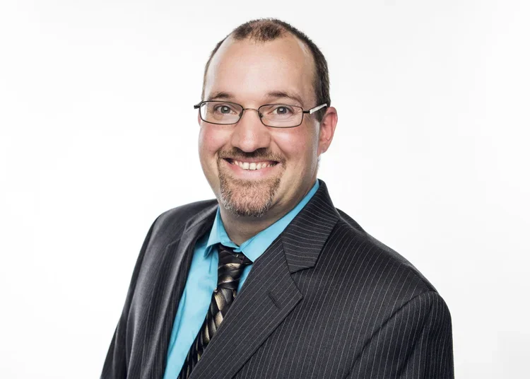 Portrait of a man with glasses, wearing a dark pinstripe suit, light blue shirt, and patterned tie, smiling against a white background.