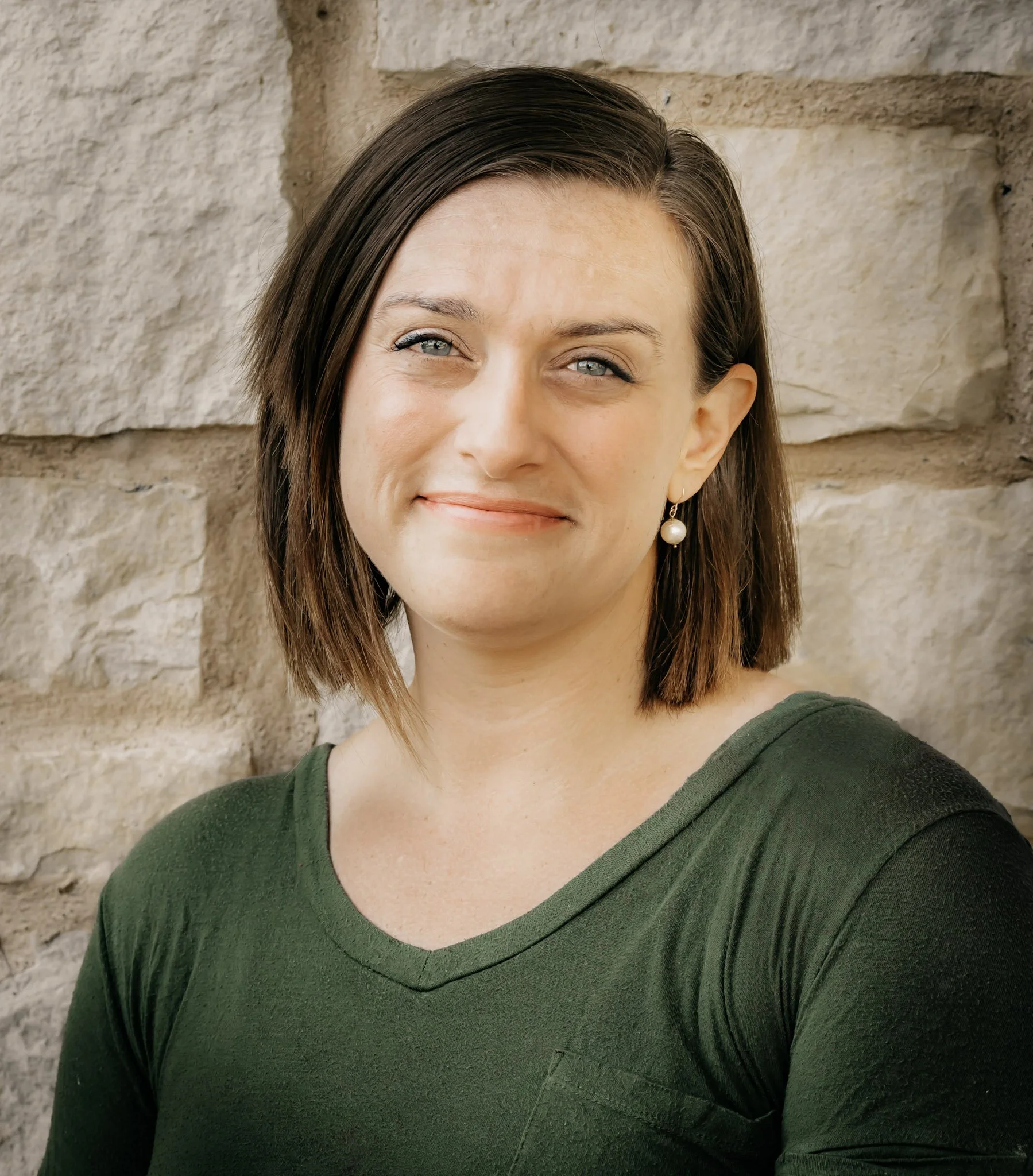 A woman with short dark hair, wearing a green shirt and pearl earrings, standing against a stone wall.