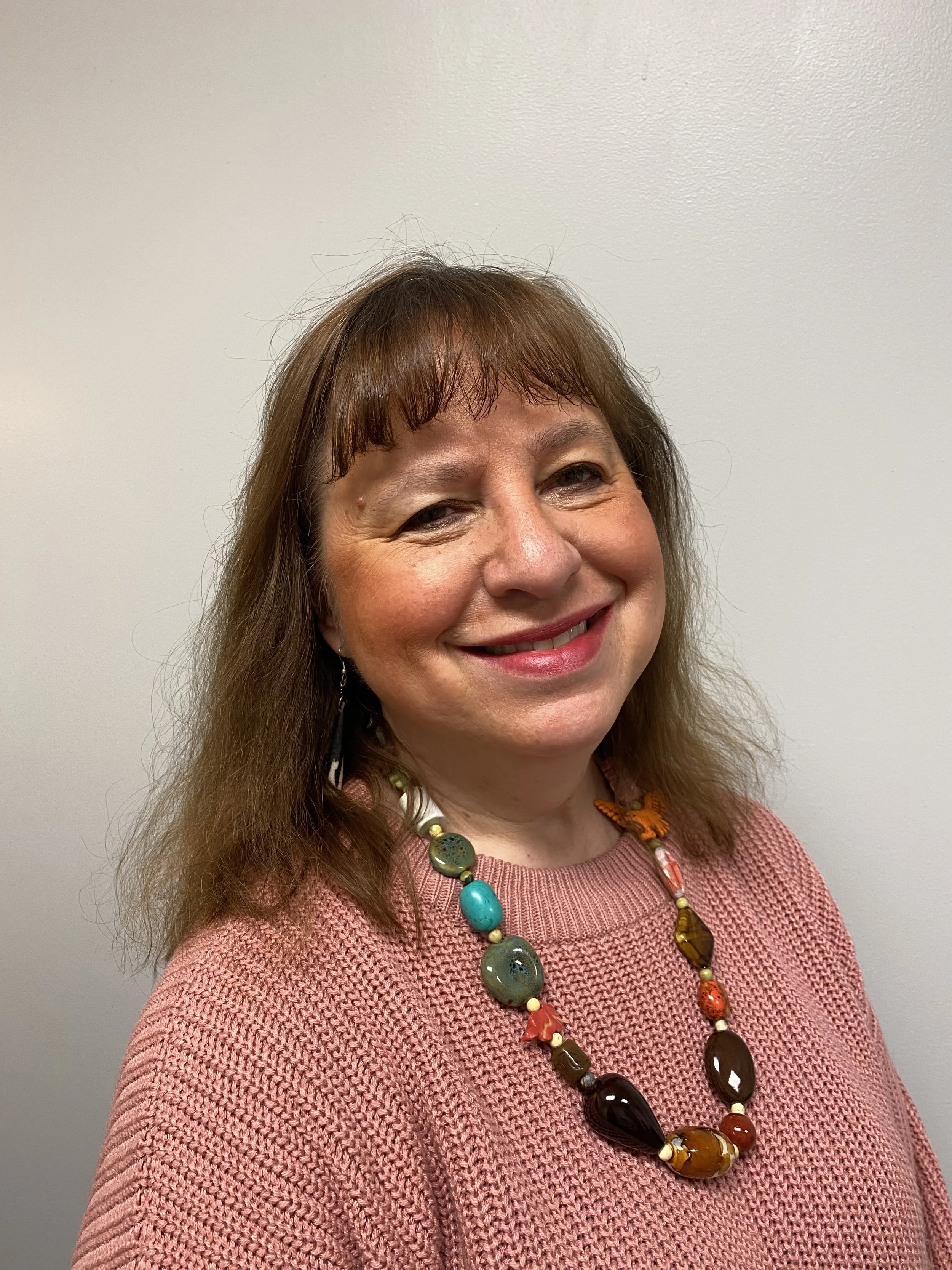 A smiling woman with shoulder-length brown hair, wearing a pink sweater and a colorful beaded necklace, standing against a plain beige wall.