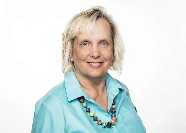 A smiling woman with short blonde hair wearing a turquoise shirt and a colorful bead necklace, standing against a plain white background.