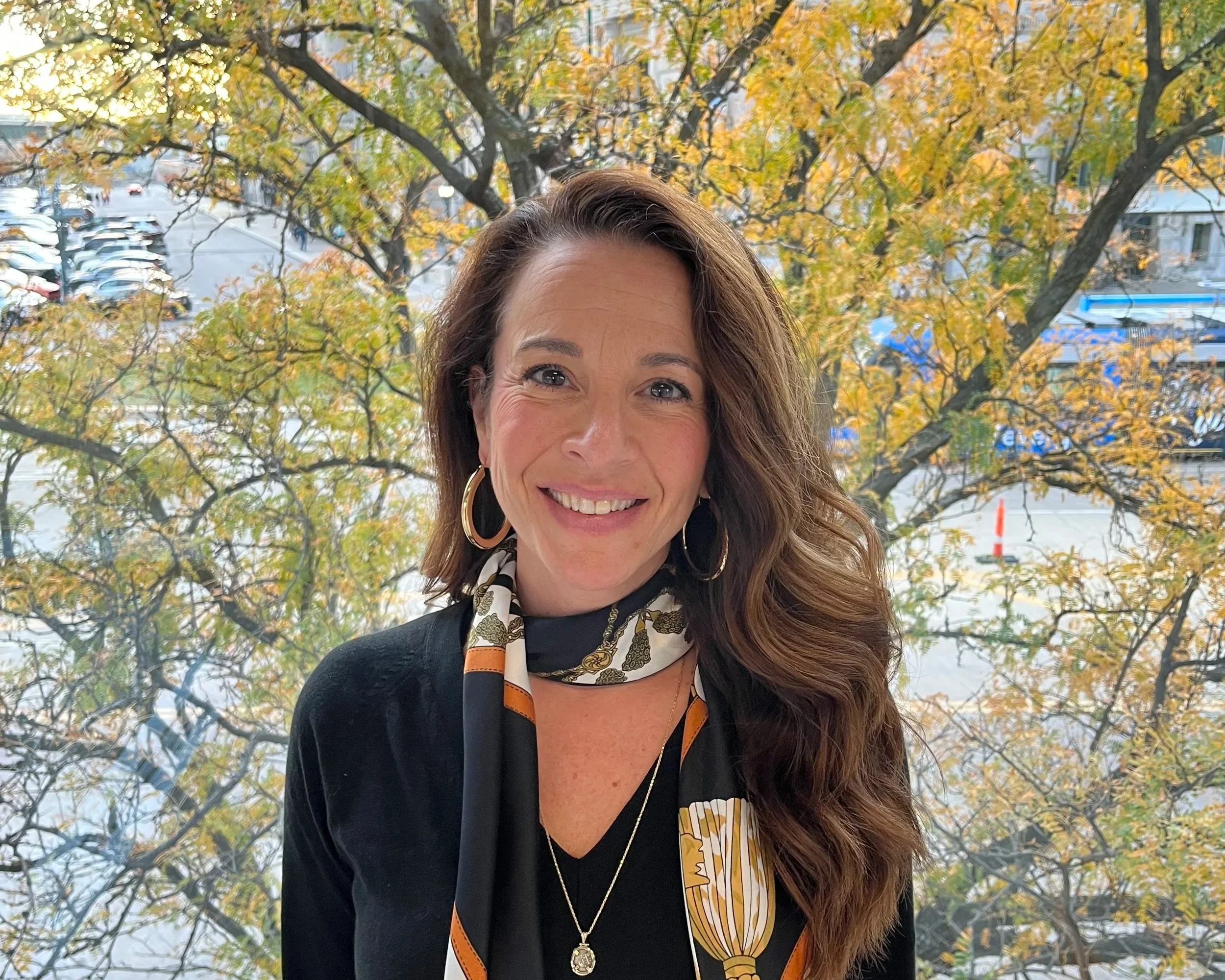 A woman with long wavy brown hair smiling at the camera standing indoors near a large window with autumn trees and a parking lot outside.