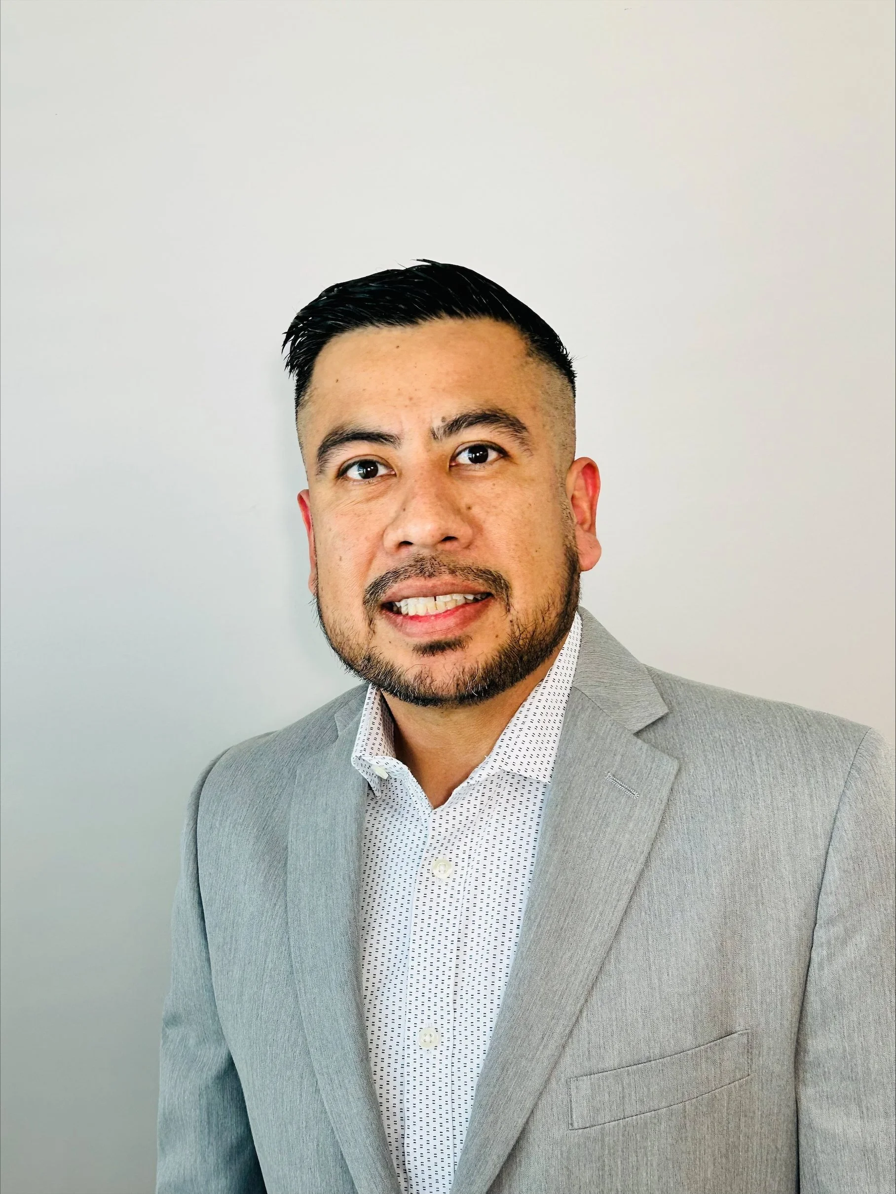 Headshot of a man with dark hair, a trimmed beard, wearing a light gray suit and a white dress shirt with small black dots, standing against a plain light-colored background.