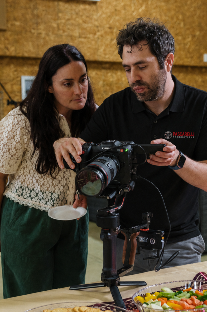 A man and woman looking at a Canon camera mounted on a tripod, with food items on the table in front of them.