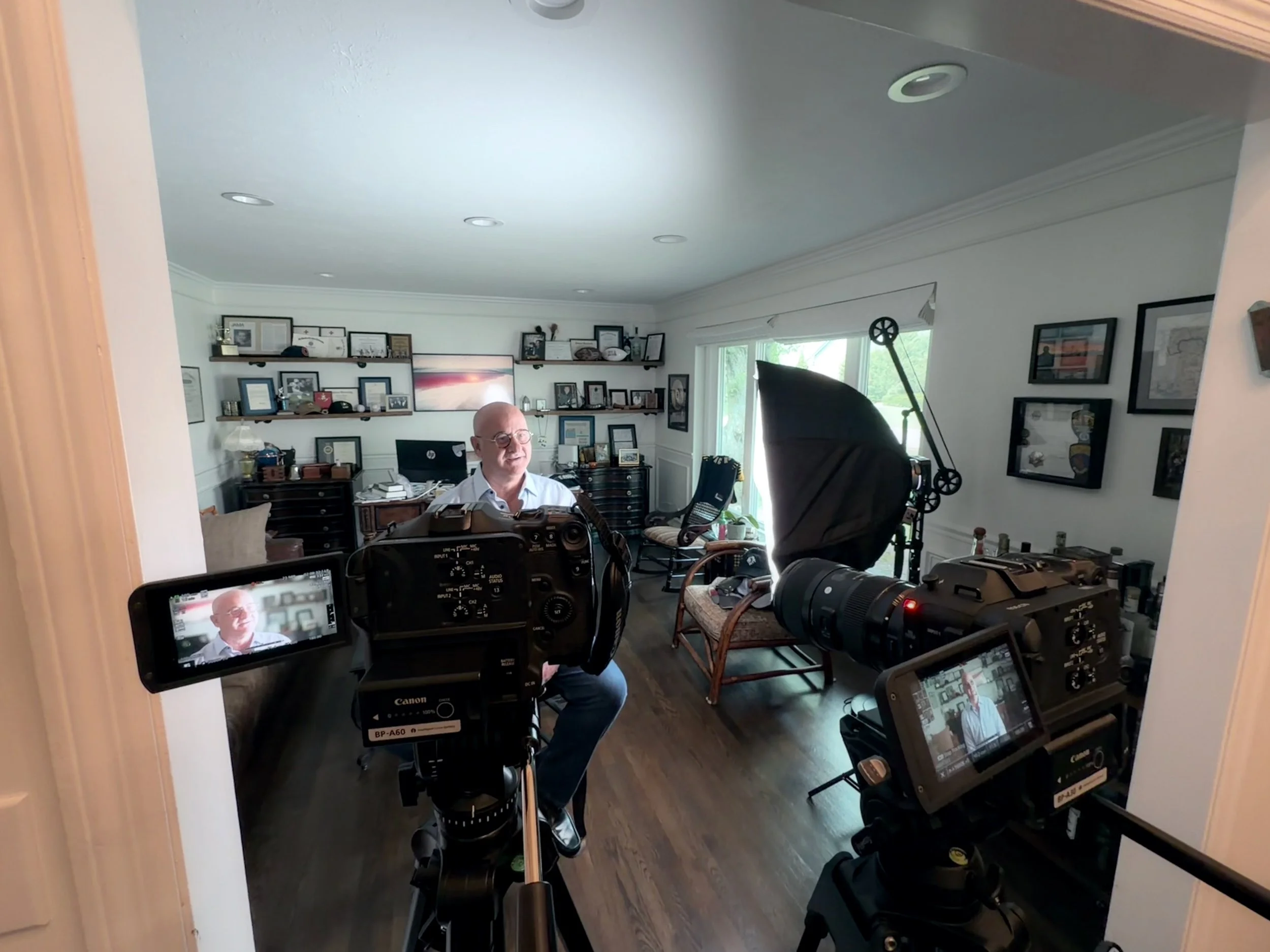 Inside a home recording studio with a man sitting in front of a camera, surrounded by photography and filming equipment, including cameras, lights, and monitors. The room has framed pictures and shelves on the walls, a large window with natural light, and various chairs and furniture.