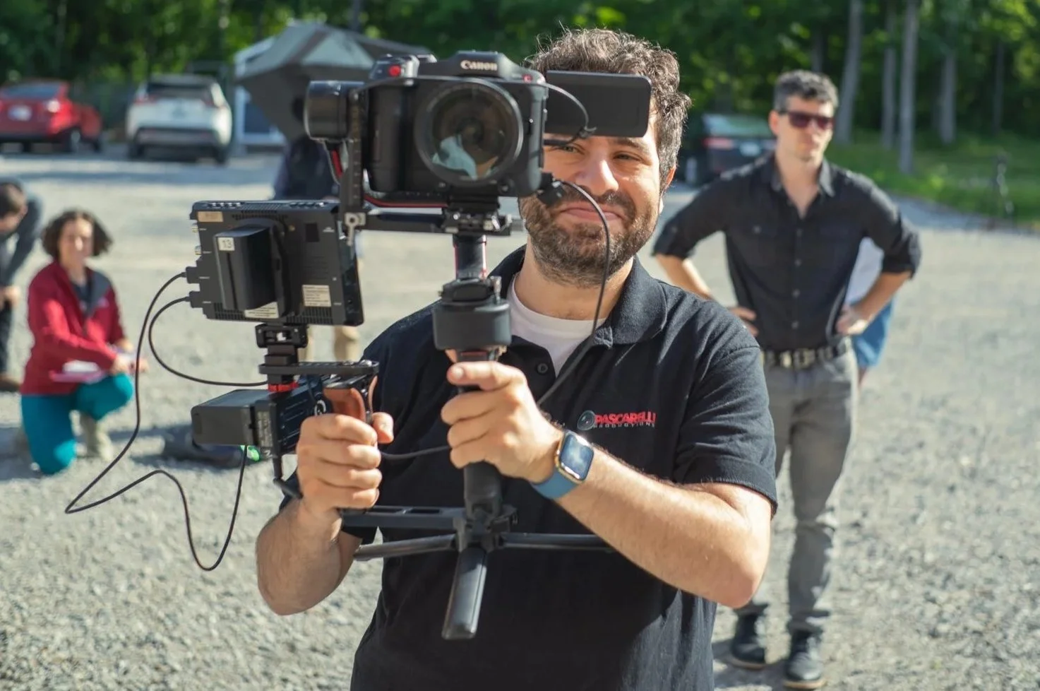 Man operating a professional camera on a stabilizer during a film shoot outdoors with a group of people in the background.