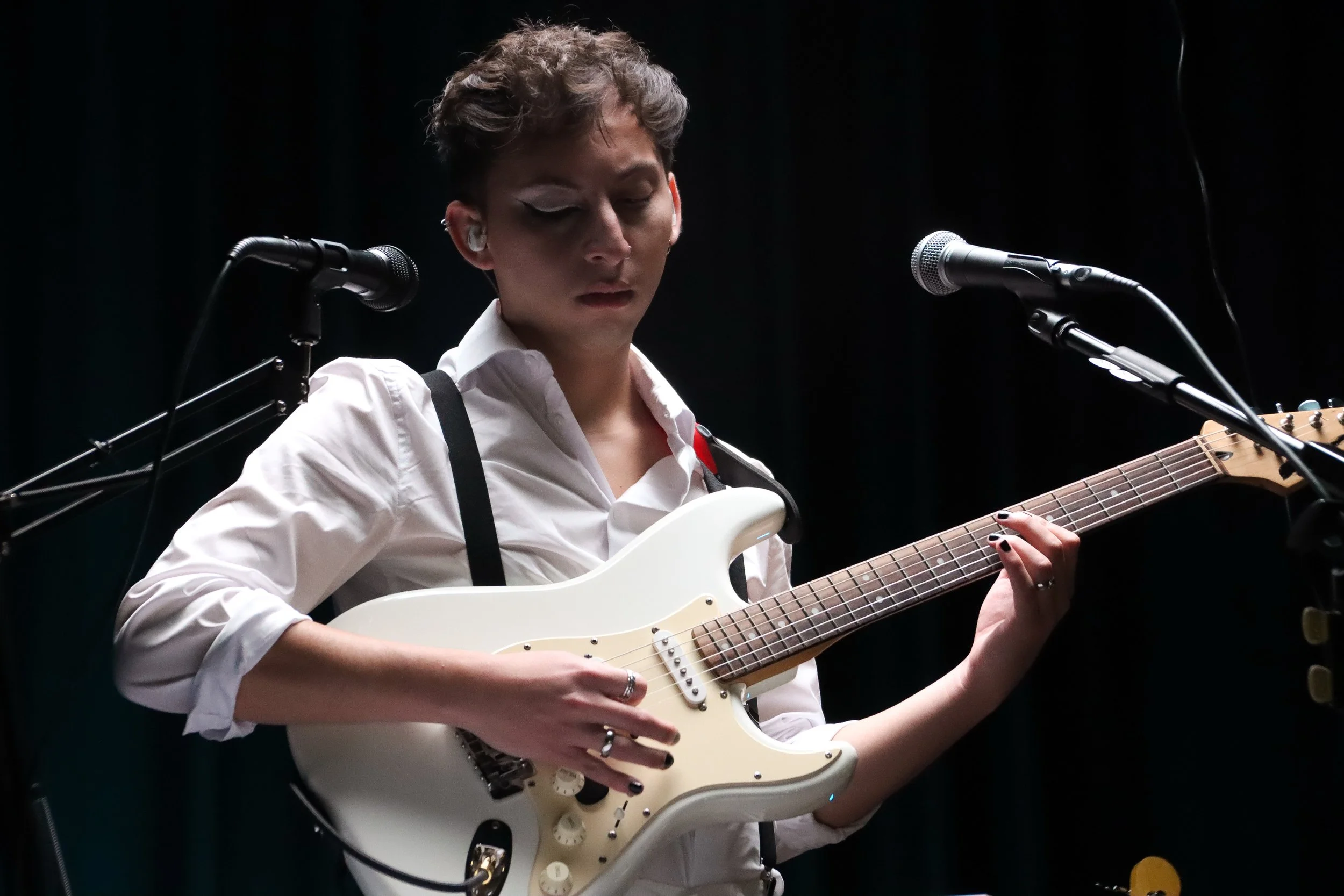 Persona tocando guitarra eléctrica en un escenario con micrófonos y fondo oscuro