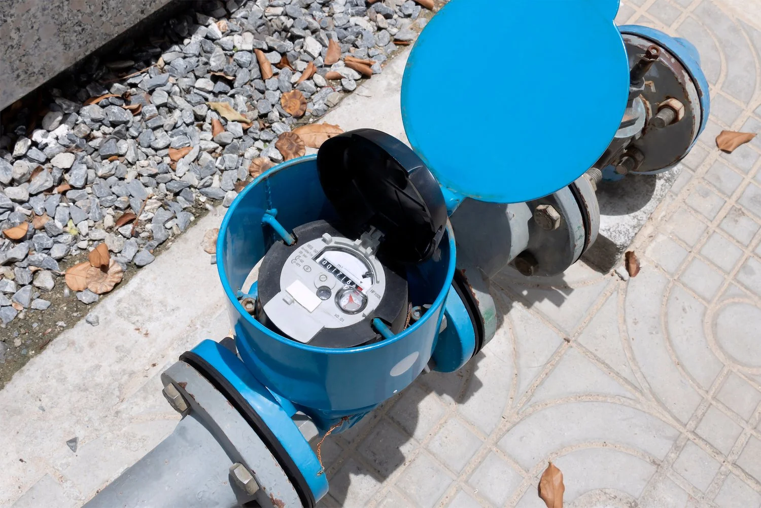 An outdoor water meter with a blue cover and a black dial, connected to a pipe, situated between a gravel area and a tiled pavement with fallen leaves.
