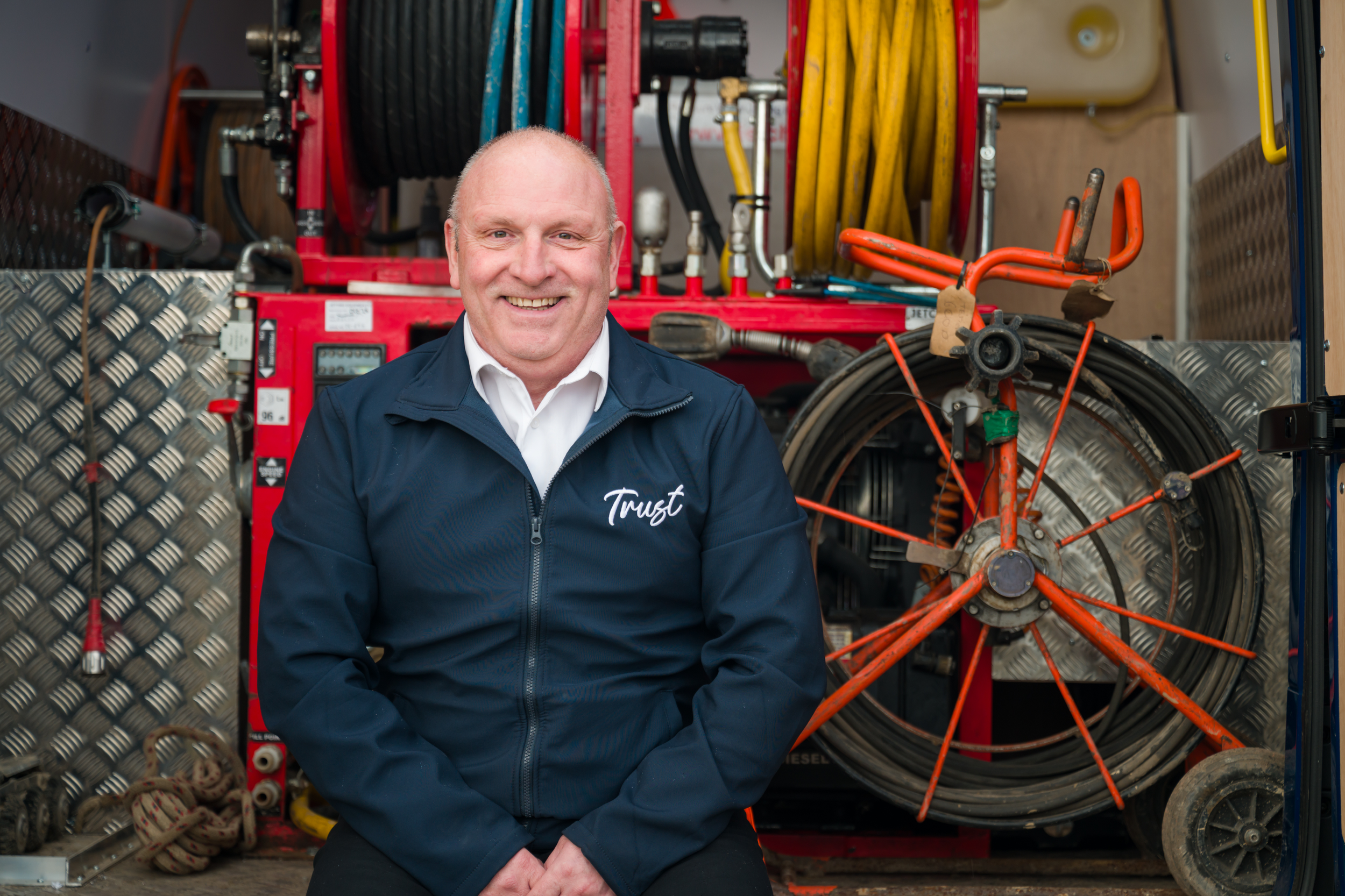 Male worker in blue Trust branded jacket sat in from of drainage equipment