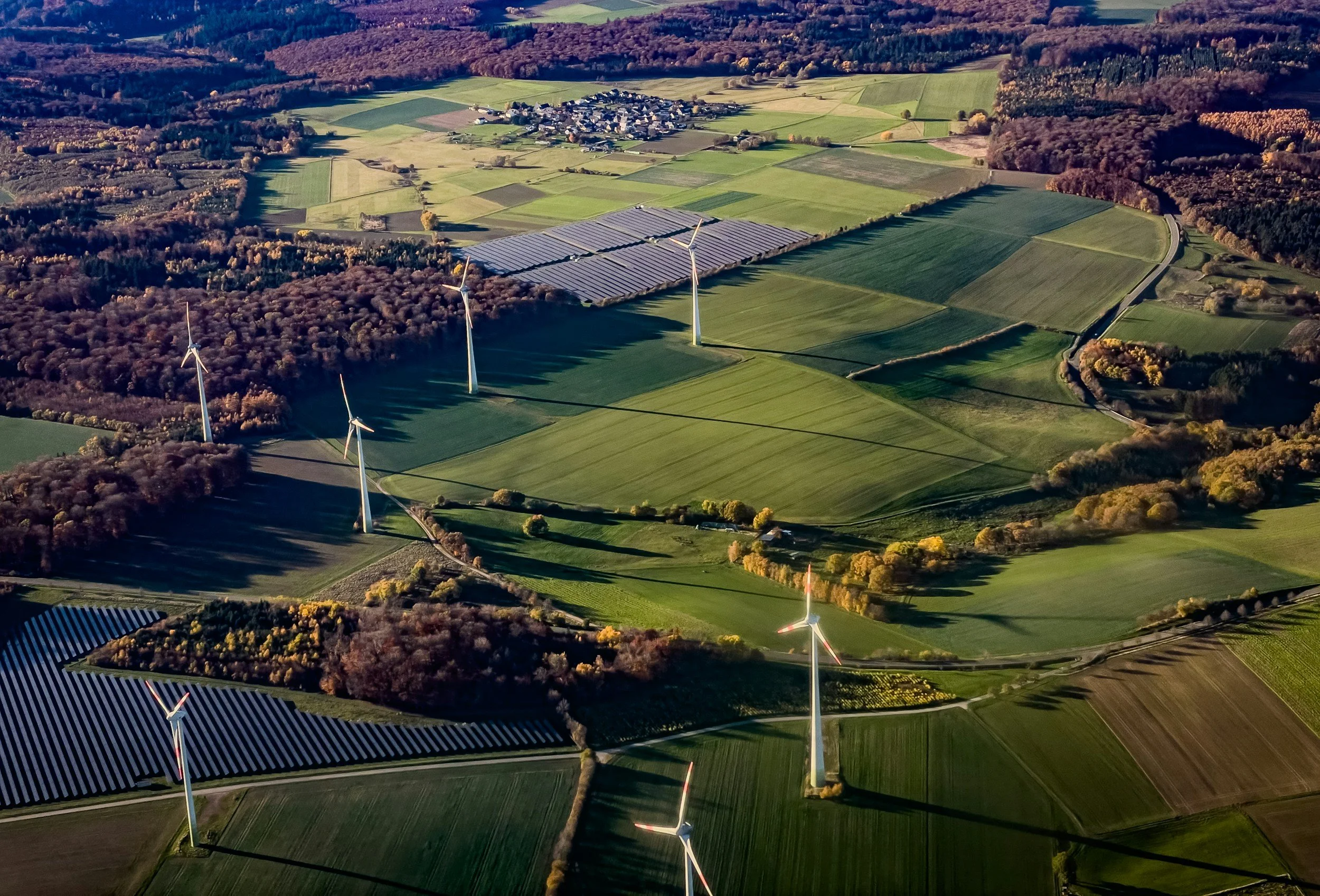 Aerial view of a rural landscape featuring wind turbines among green fields, patches of woodland, and a small farm or village in the distance.