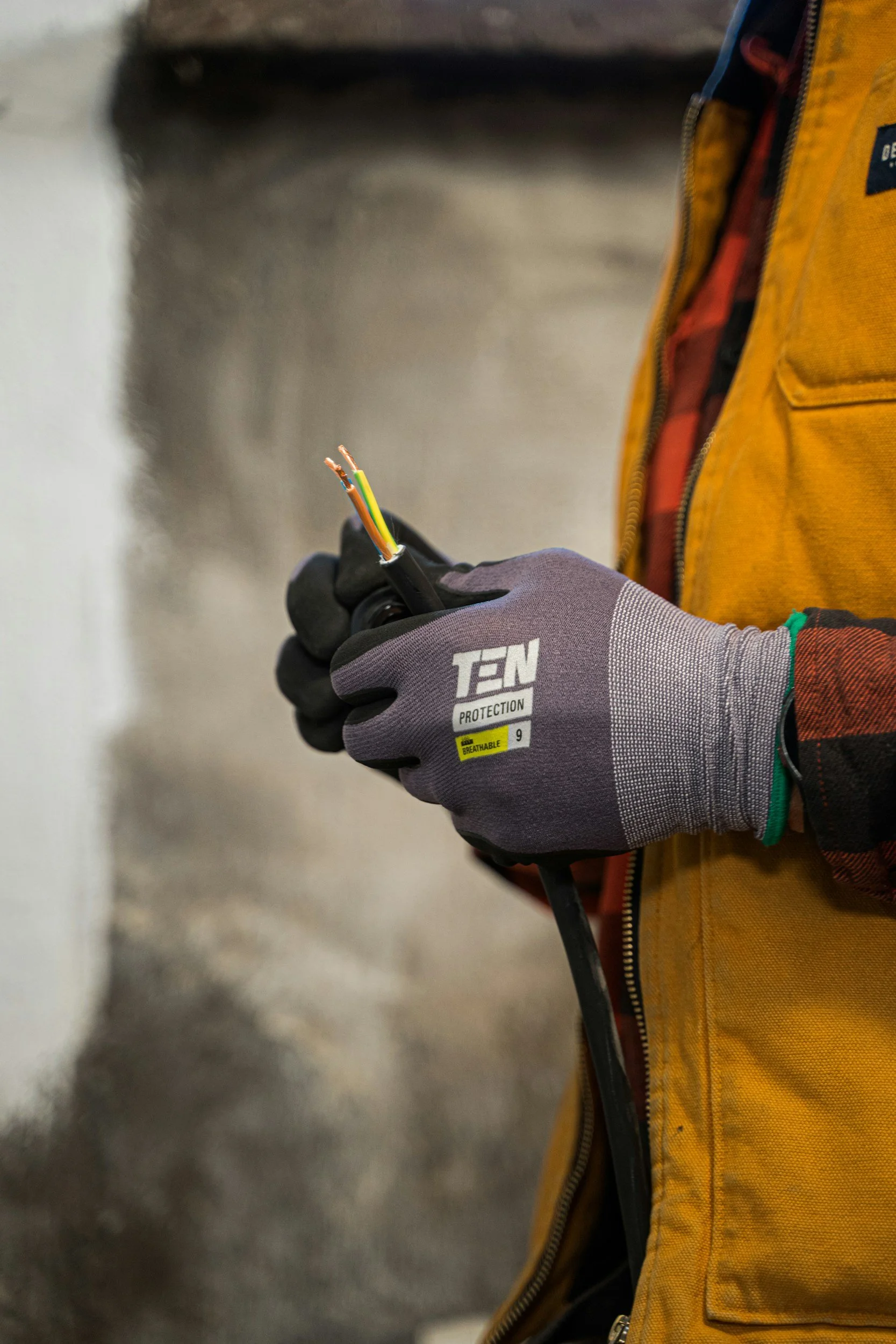 A person wearing gray gloves labeled 'TEN' and a yellow work jacket is holding and inspecting a black electrical cable with colored wires exposed.