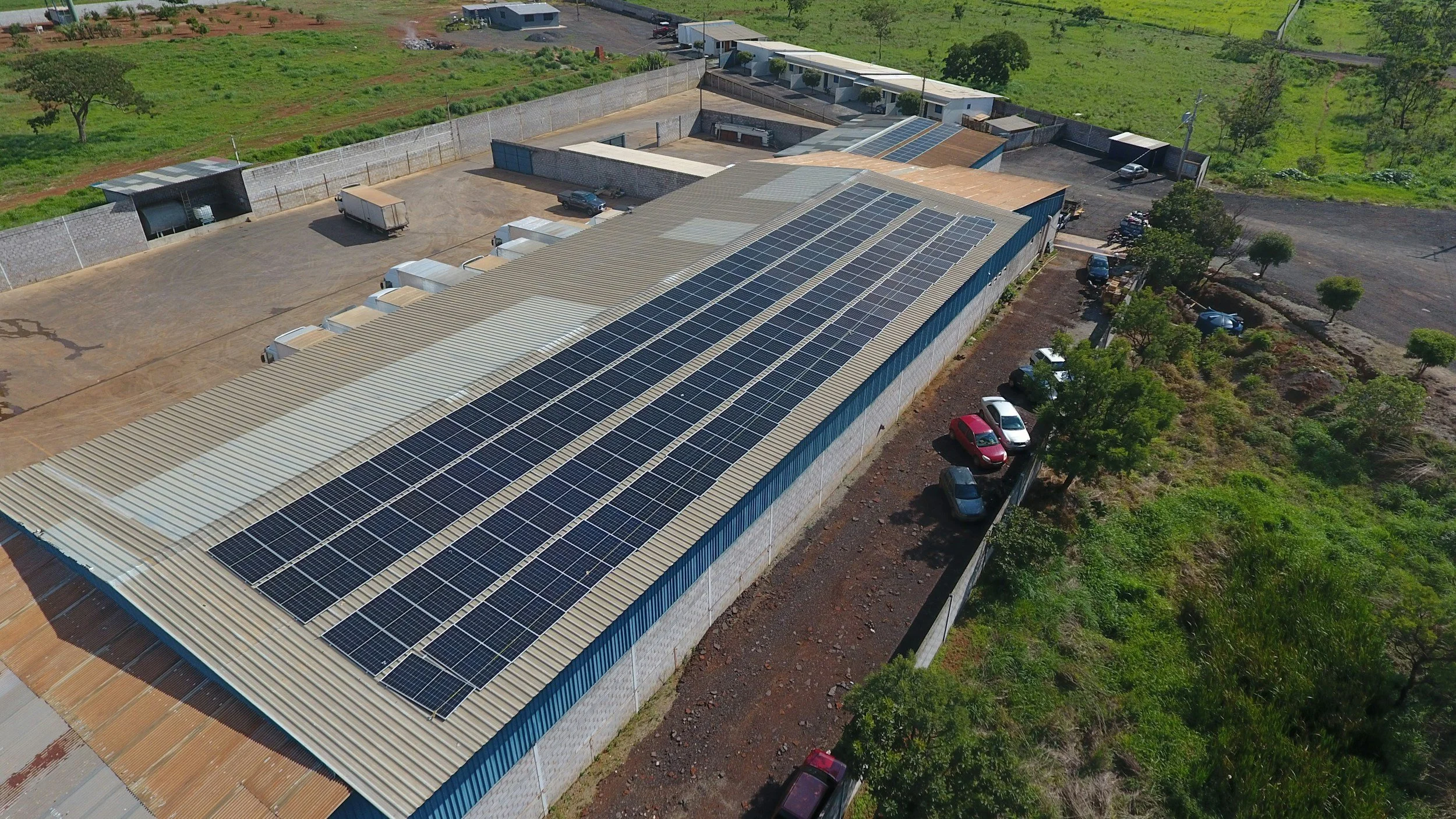 Aerial view of a large industrial building with numerous solar panels on the roof, parked cars along one side, surrounded by greenery and open land.