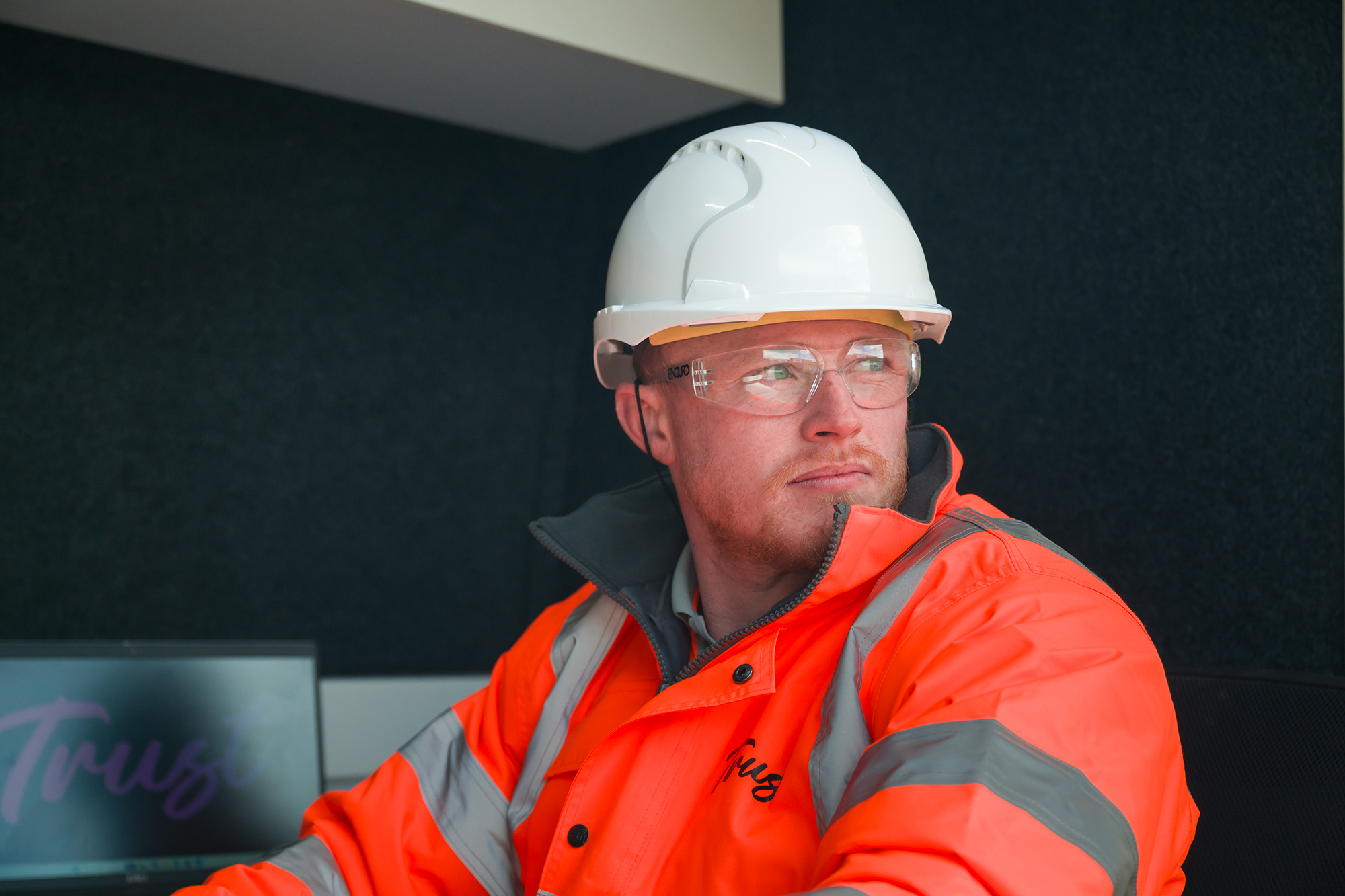 Male Trust worker wearing a white hard hat, safety glasses and orange hi-vis clothing looking slightly away from the camera on a water job site.